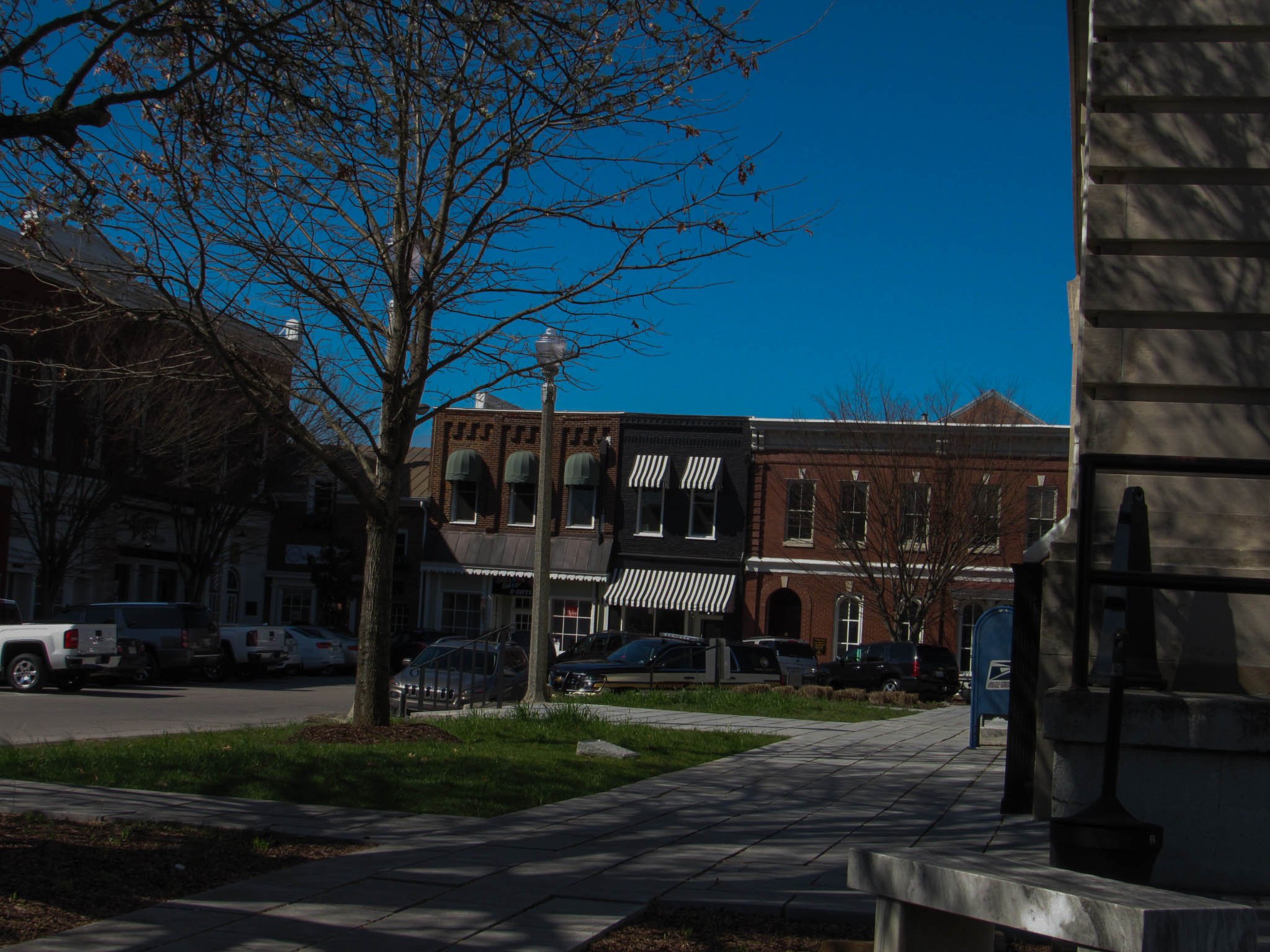 A small downtown street scene with brick buildings, parked cars, leafless trees, a sidewalk, a lamppost, and a mailbox under a clear blue sky.