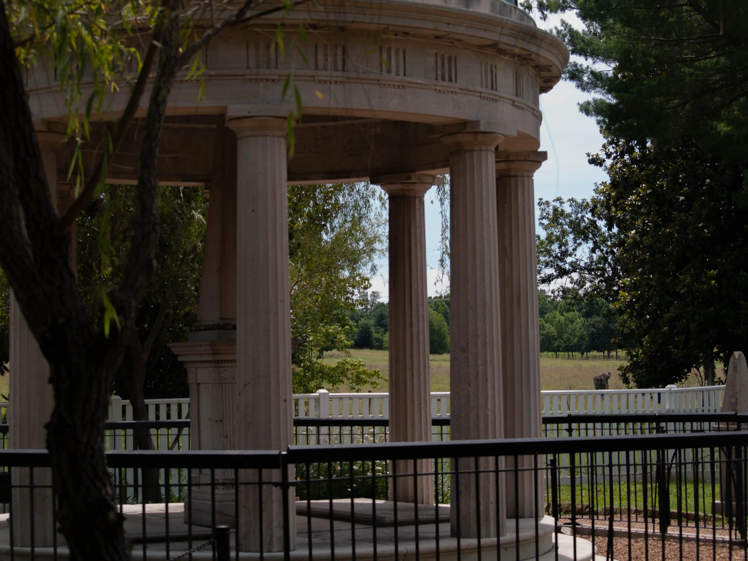Outdoor view of a historic marble bandstand with fluted columns, surrounded by trees and a white fence, in a park or garden setting.