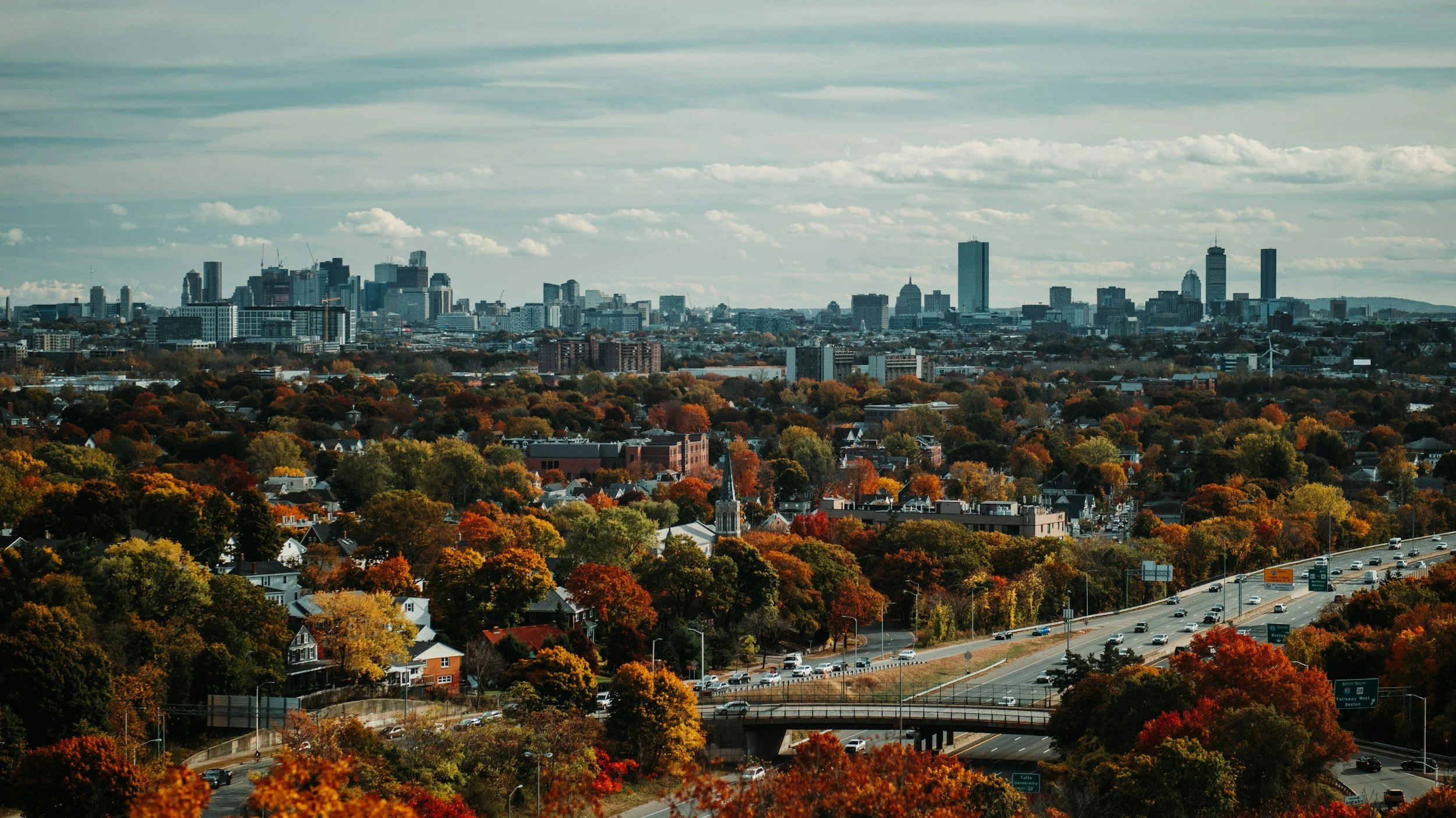 A city skyline in the distance with a suburban neighborhood in the foreground, showing trees with autumn foliage and a busy highway.