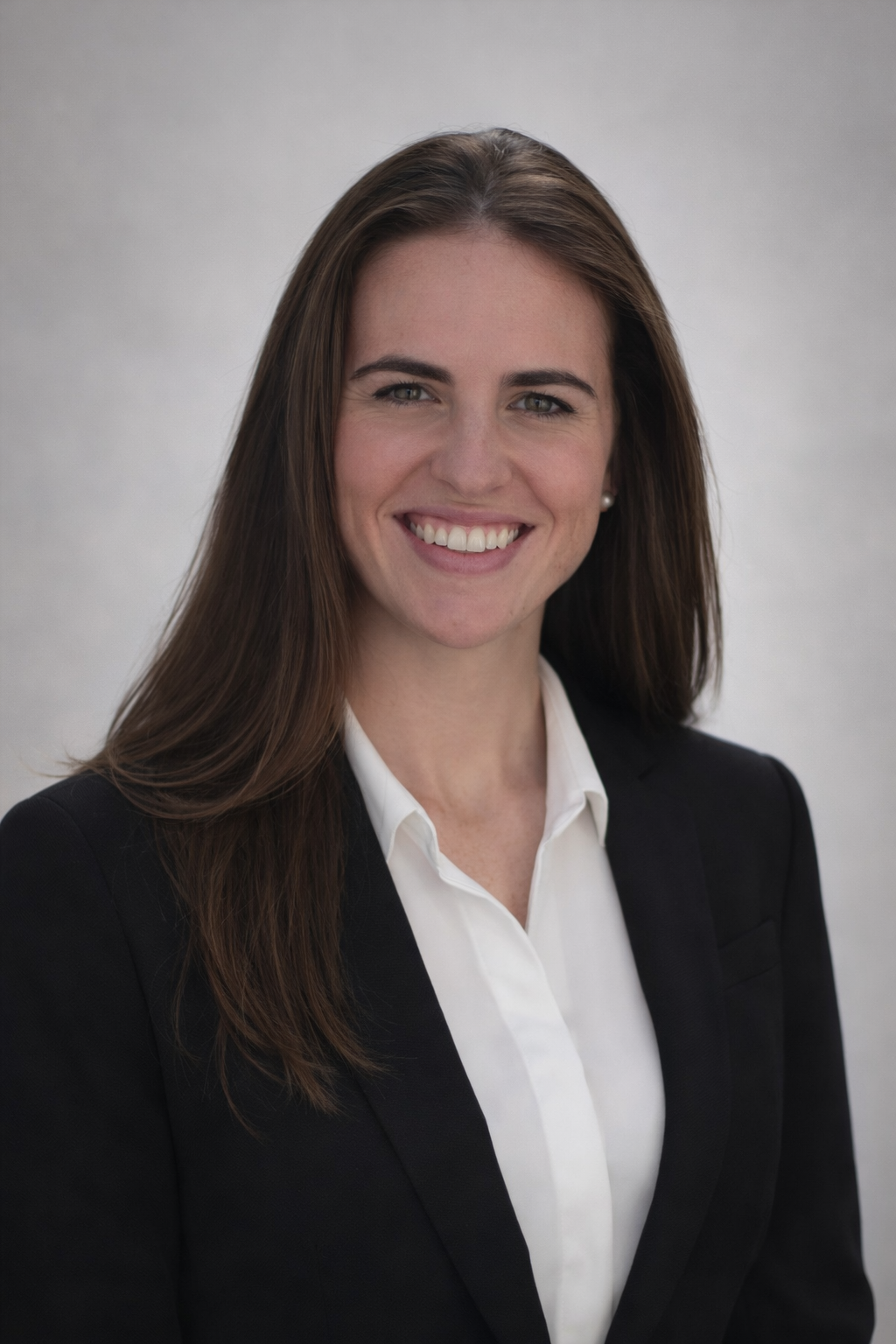 A professional woman with long brown hair, wearing a black blazer and white shirt, smiling at the camera against a plain light gray background.