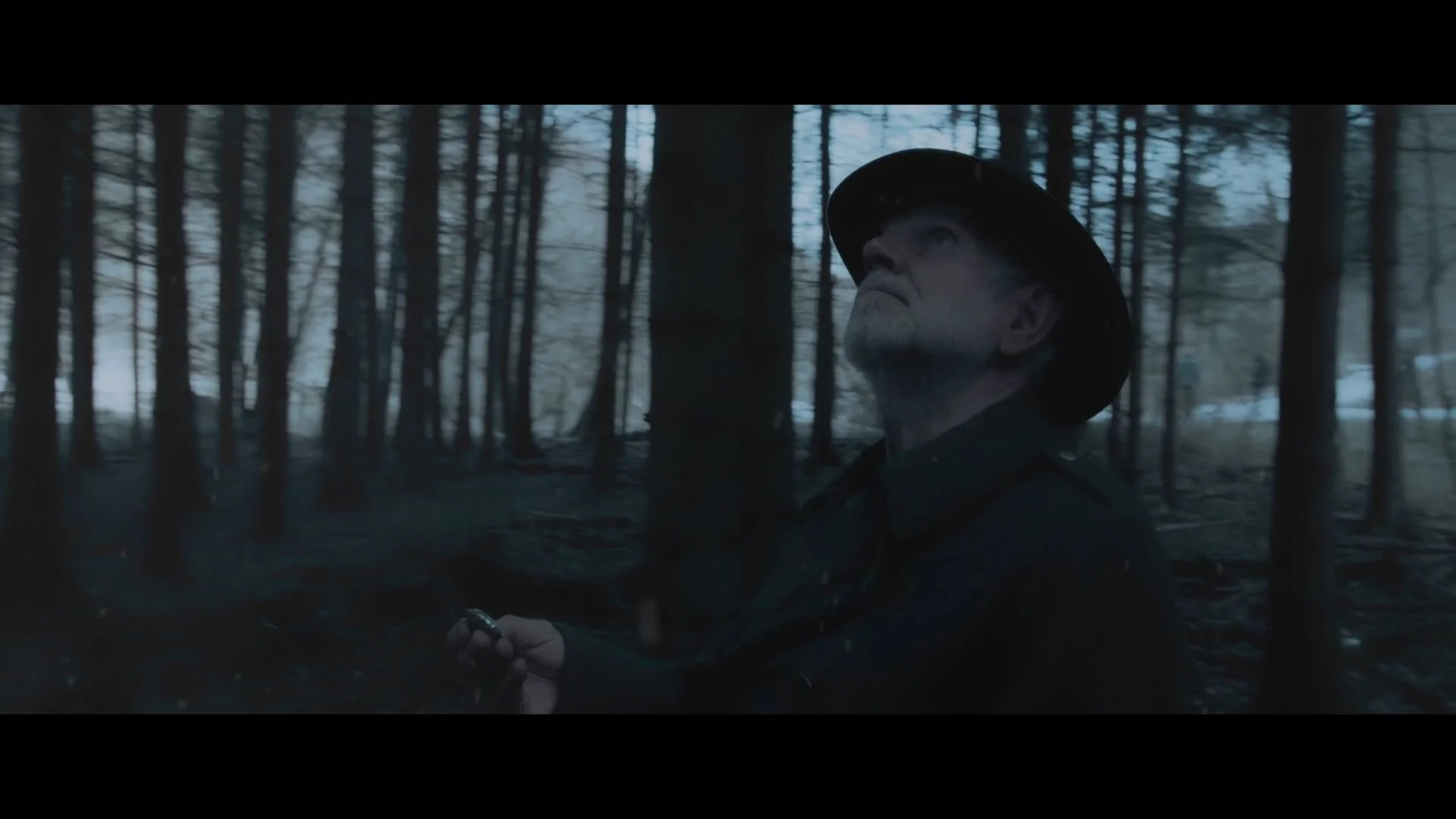 Ein älterer Mann mit Hut steht im dunklen Wald und sieht nach oben, während er ein Gerät in der Hand hält.