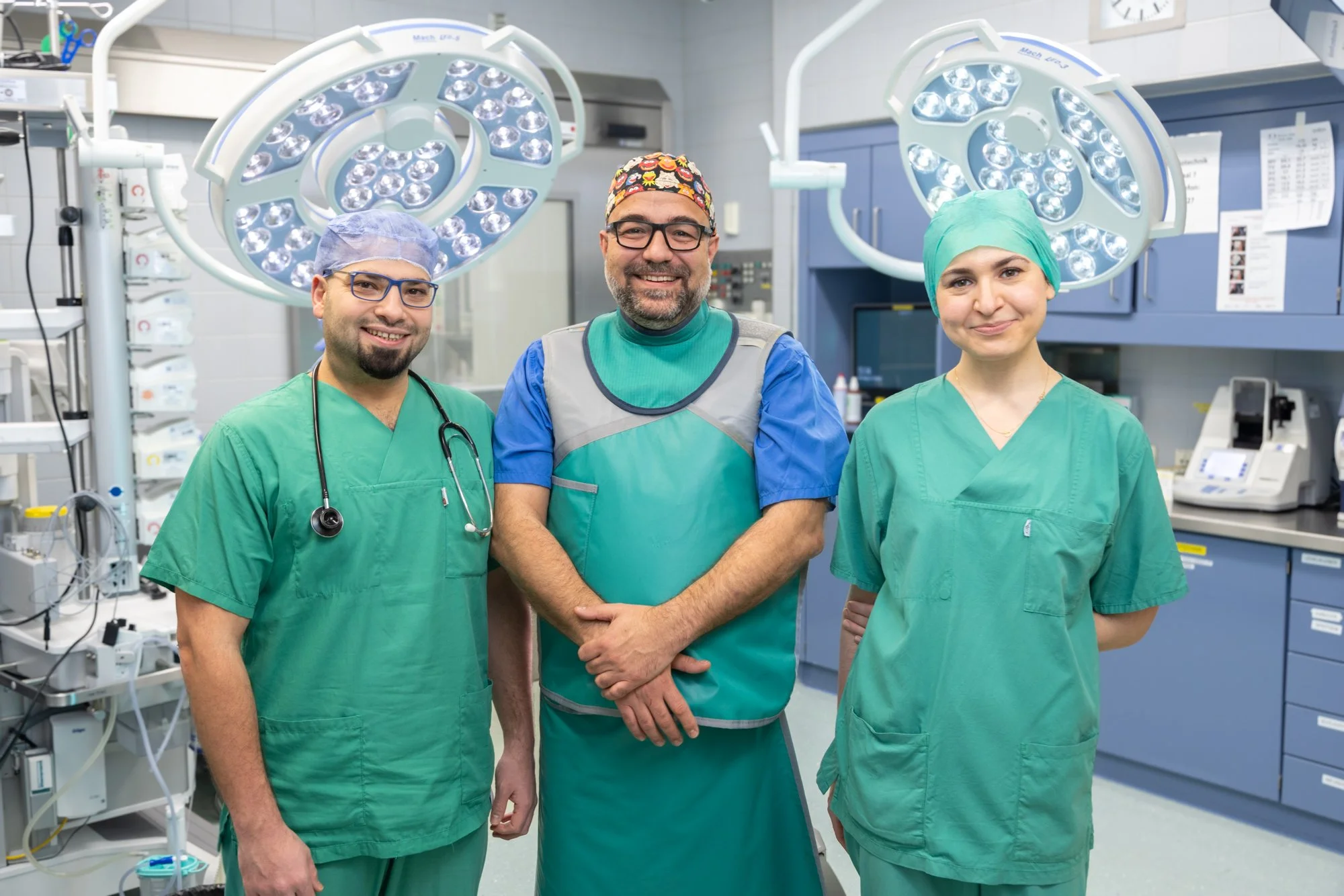 Drei medizinisches Personal in Operationssaal, zwei Männer und eine Frau, alle tragen grüne OP-Kleidung, mit OP-Lampen und medizinischer Ausrüstung im Hintergrund.