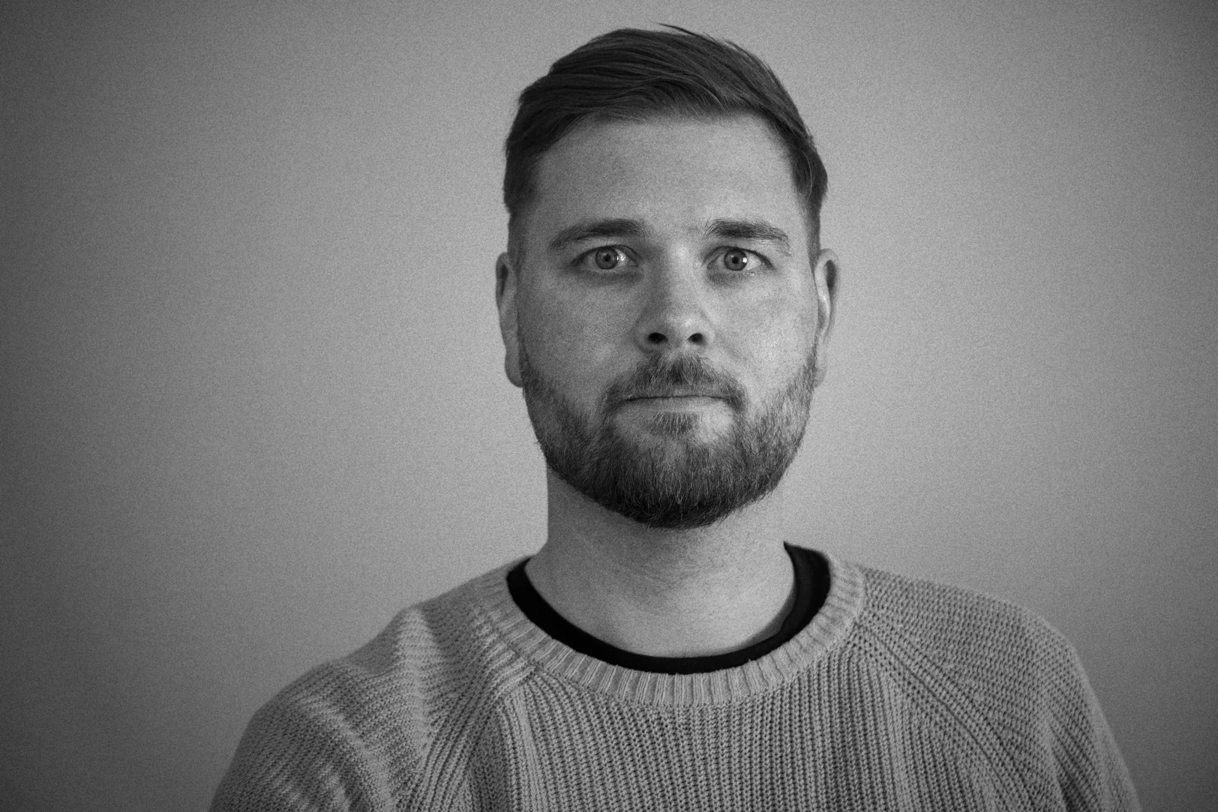 Schwarz-weiß Portrait eines Mannes mit kurzem Haar, Bart und casual Pullover vor neutralem Hintergrund.