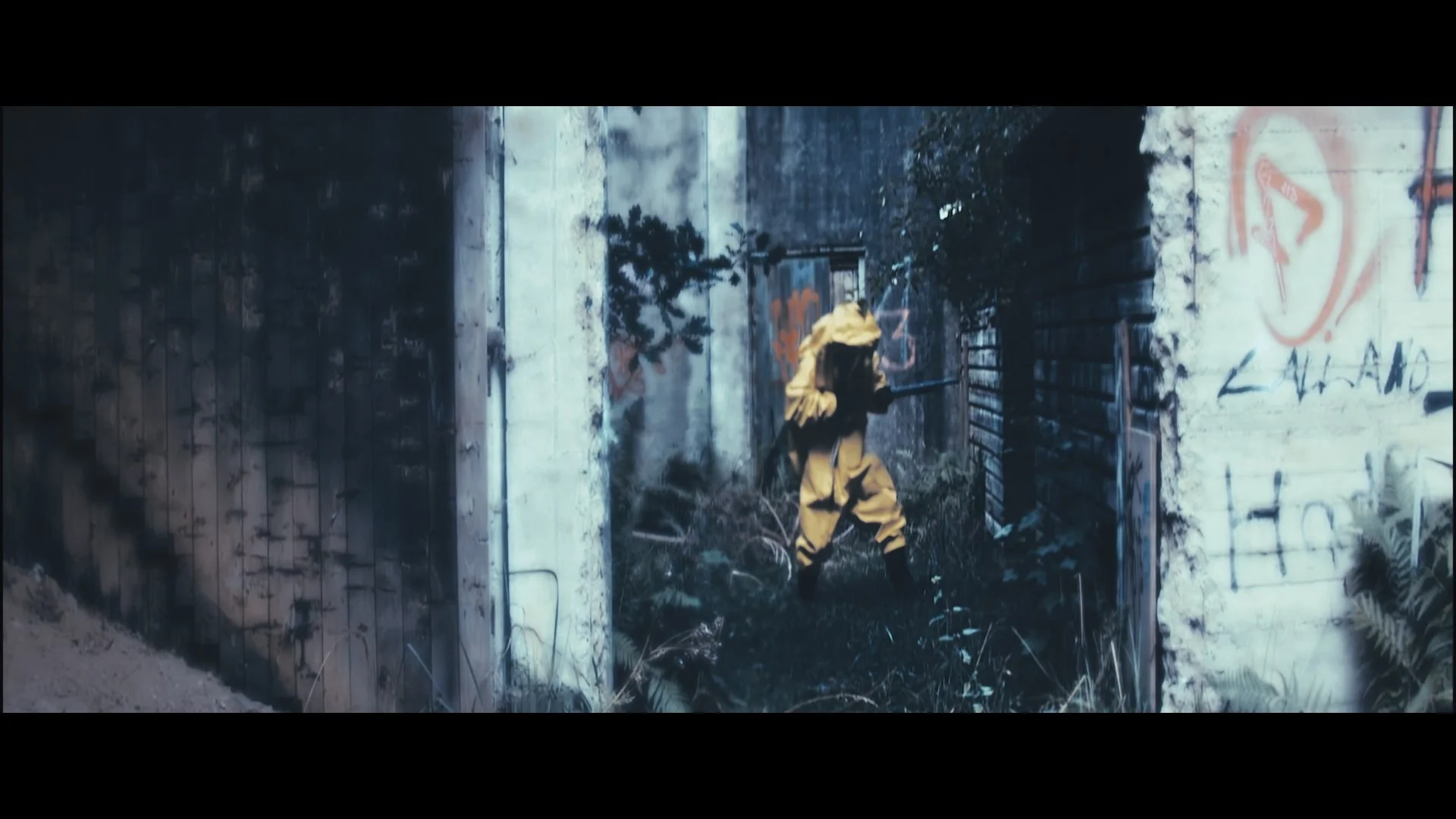 Person in gelber Schutzkleidung und Atemschutzmaske in einer verlassenen, mit Graffiti besprühten urbane Umgebung mit verfallenem Mauerwerk.