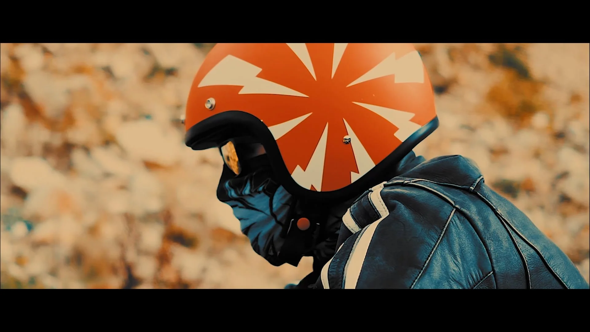 Ein Mensch trägt einen orangefarbenen Motorradhelm mit weißen Mustern, eine schwarze Uhr und eine schwarze Jacke mit weißen Akzenten, im Hintergrund liegen viele Herbstblätter.