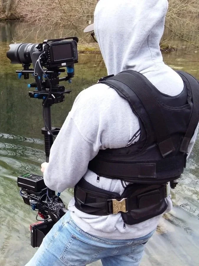 Person in Hoodie und Weste mit Kamera in der Hand am Fluss