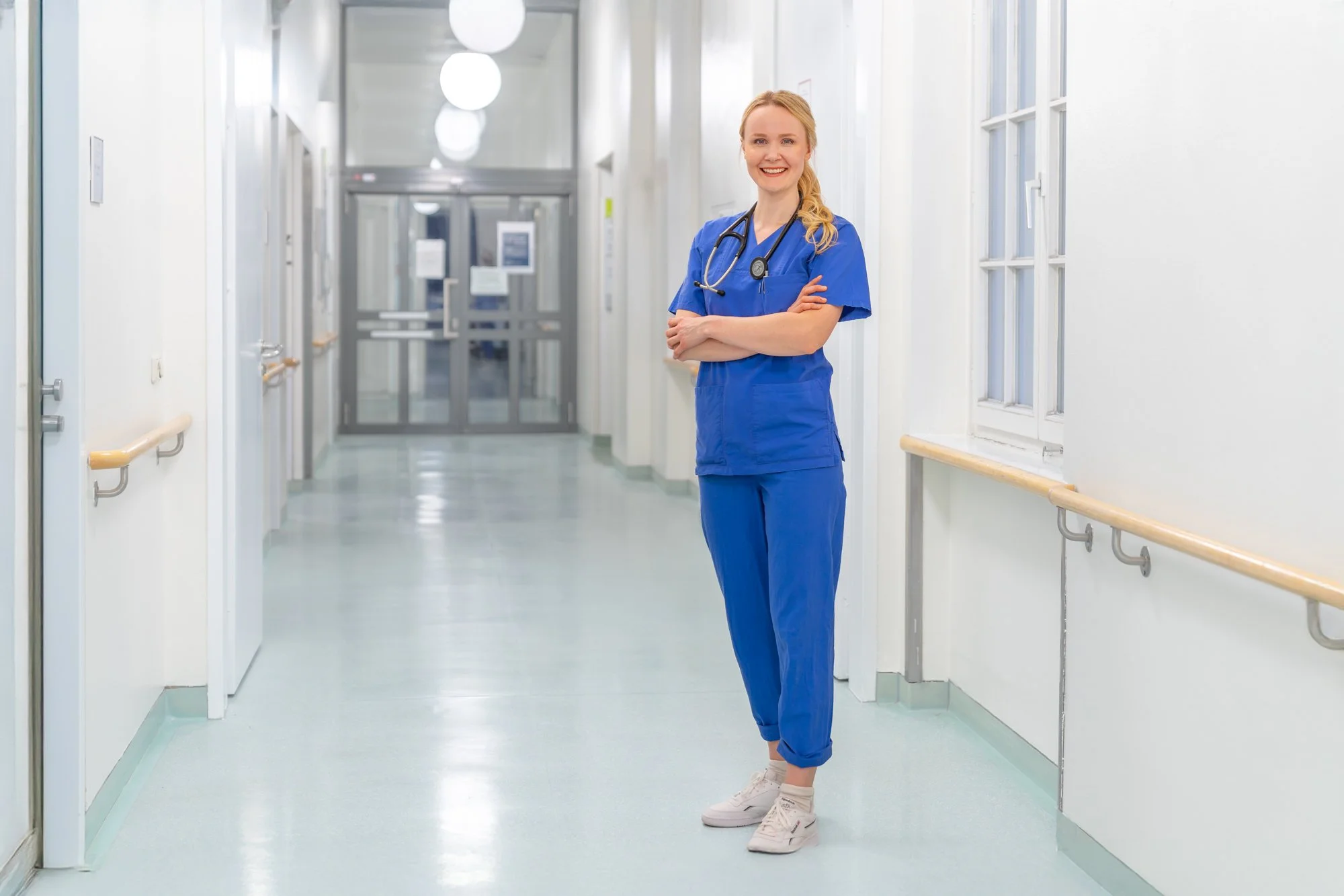 Junge weibliche Krankenschwester oder Pflegekraft in blauer Berufskleidung in einem Krankenhausflur, lächelnd mit Stethoskop um den Hals, Arme verschränkt.