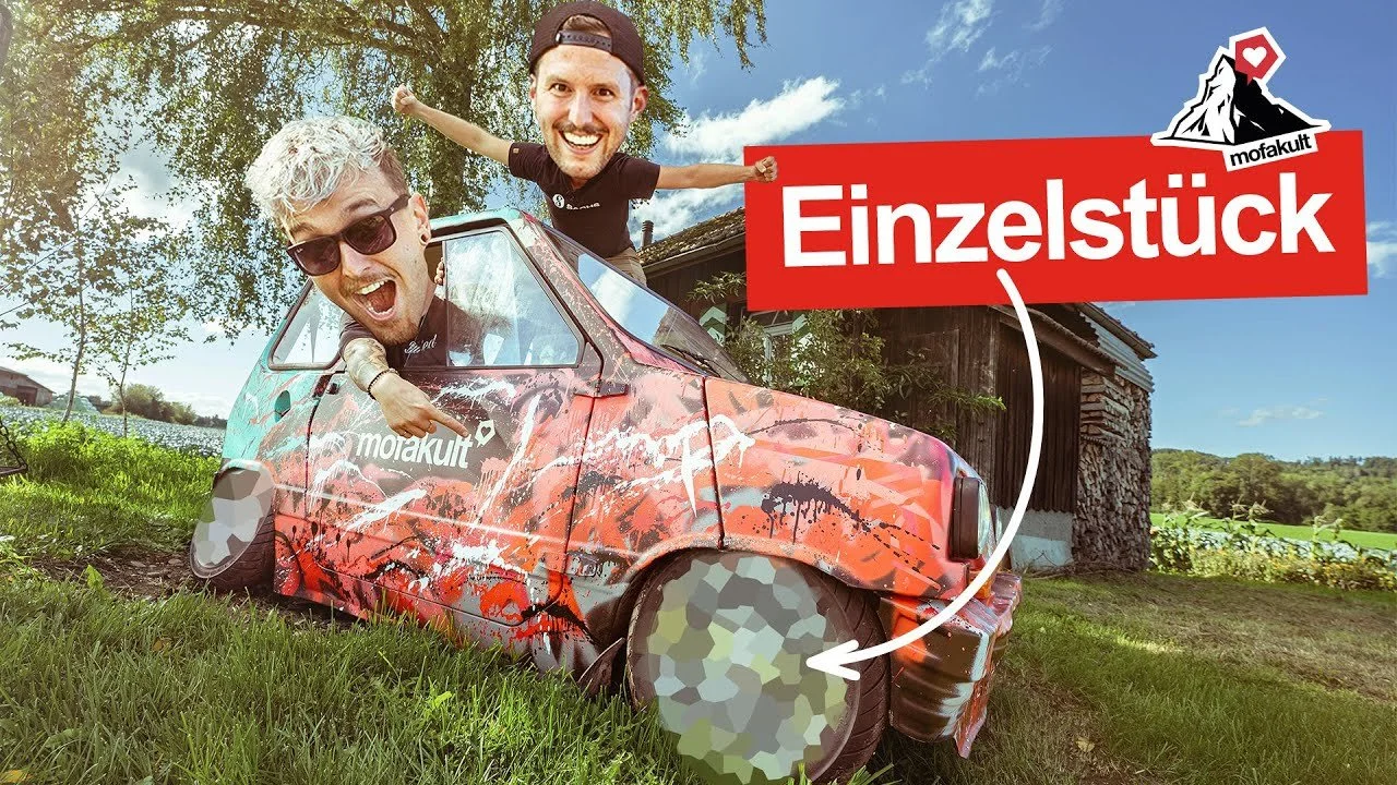 Zwei Männer mit übergroßen Köpfen posieren neben einem lackierten, kleinen Auto. Eines der Männer lehnt auf dem Auto, während der andere auf dem Dach steht. Das Bild enthält einen Textblock mit der Aufschrift 'Einzelstück' und ein Logo mit einem Berg und Herz.