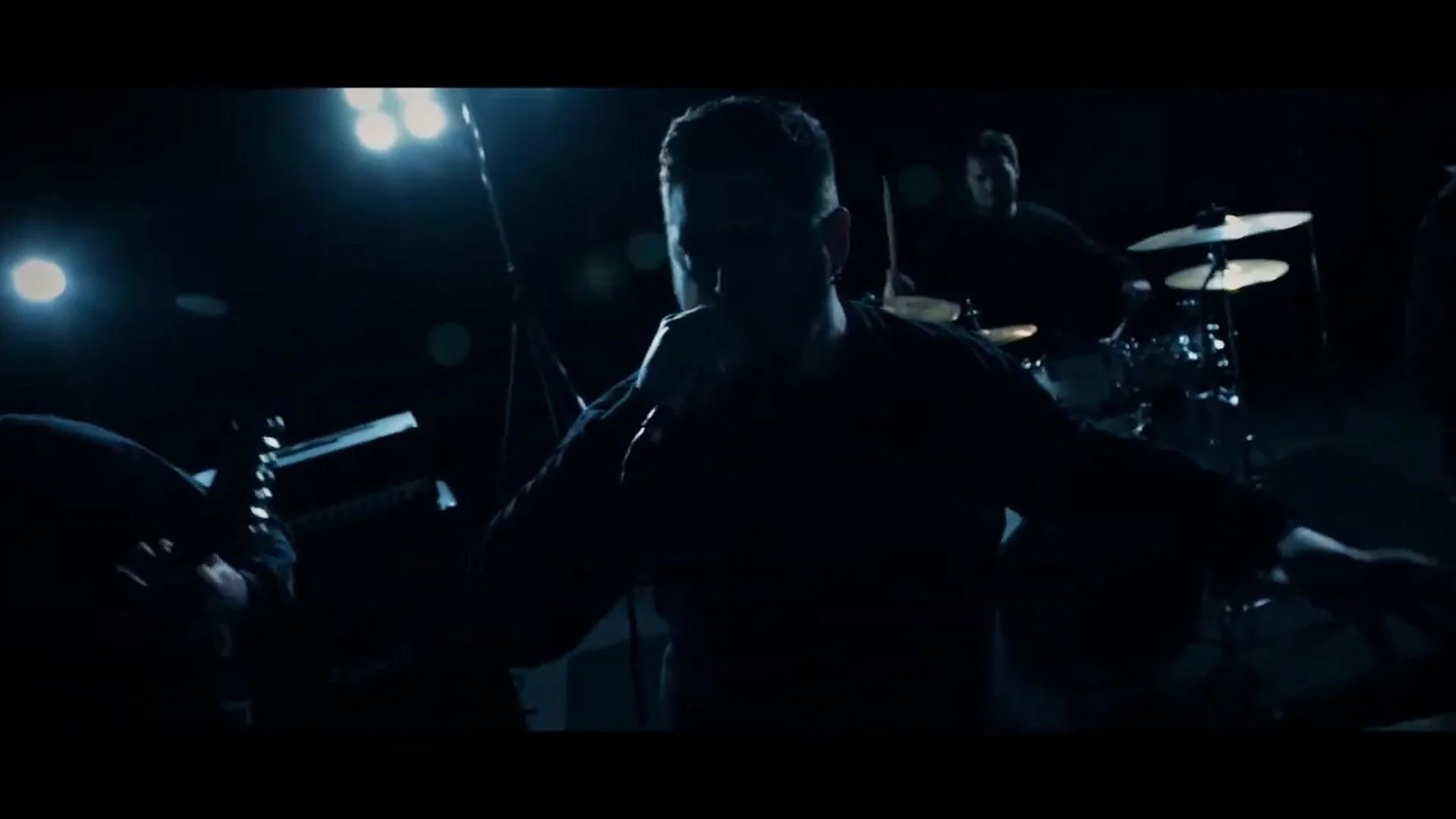 Dunkle Aufnahmeposition einer Band mit Mitgliedern, die auf einer Bühne spielen, im Hintergrund und im Schatten beleuchtete Schlagzeug- und Keyboard-Setups, mit starken Lichtquellen hinter den Musikern.