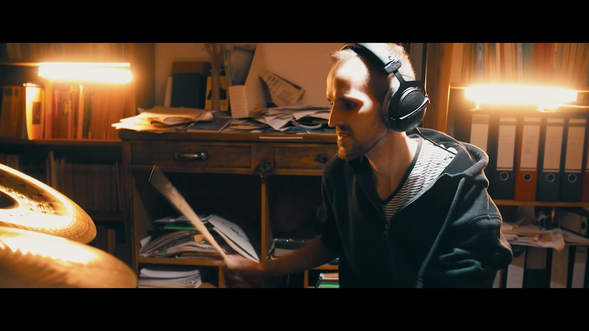 Ein junger Mann spielt Schlagzeug in einem unordentlichen Arbeitszimmer, trägt Kopfhörer und trägt dunkle Kleidung.