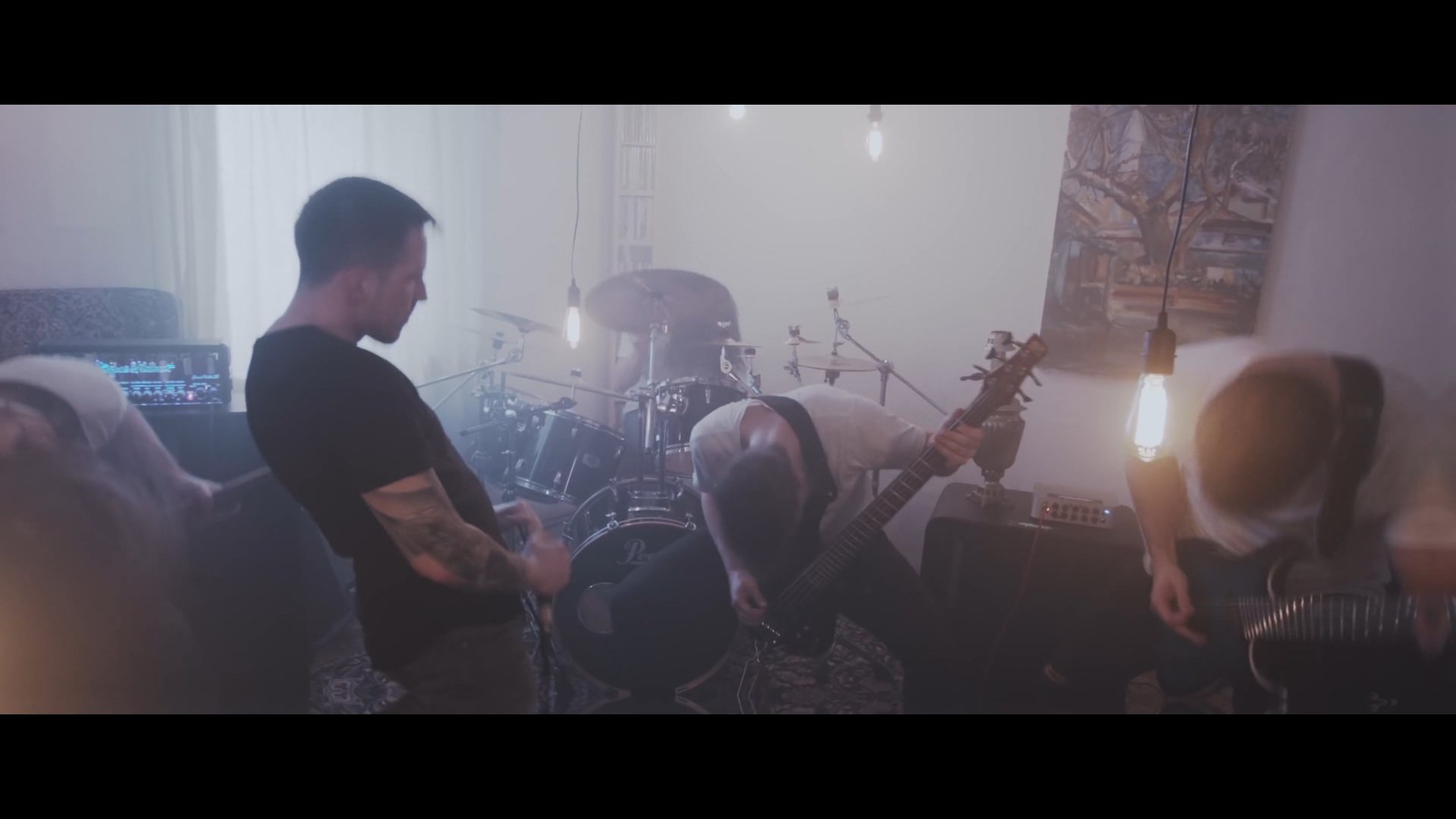 Drei Männer spielen in einem verrauchten Raum mit Musikinstrumenten, einer sitzt auf einem Sofa, die anderen auf dem Boden, mit einer Drum-Set im Hintergrund.