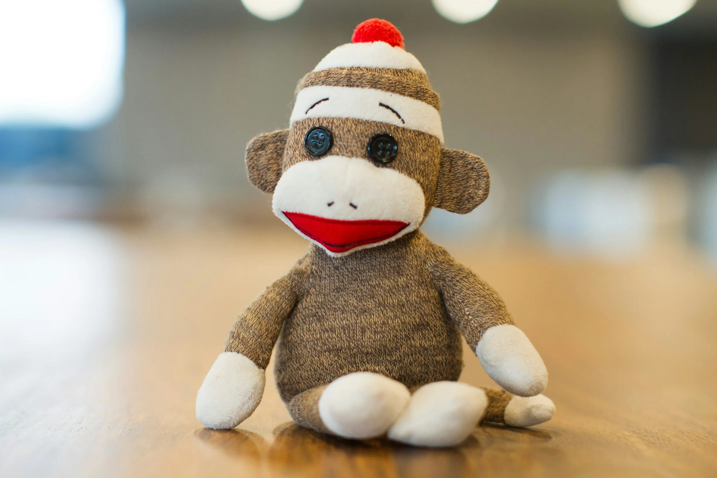 A plush sock monkey toy with fabric buttons for eyes, red and white embroidered mouth, brown sock-like body, and a Santa hat with red pom-pom, sitting on a wooden surface.