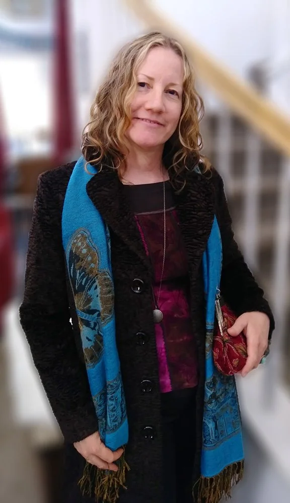 Photo of artist with wavy blonde hair and brown eyes, wearing a long coat, blue scarf and a pendant.