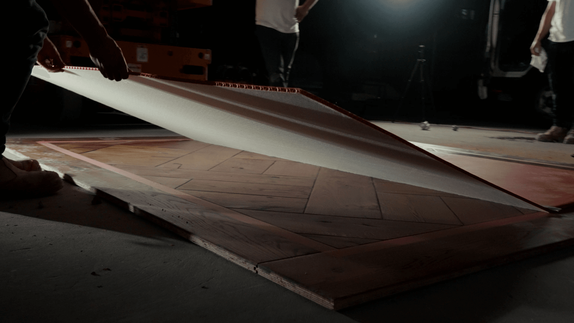 Close-up of someone unrolling a large sheet of white material over a wooden floor on a film set, with other crew members in the background.