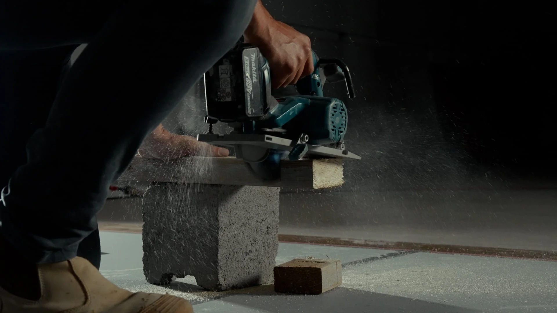 Person using a handheld power saw to cut a piece of wood on a support block, with sawdust flying, in a workshop or construction setting.