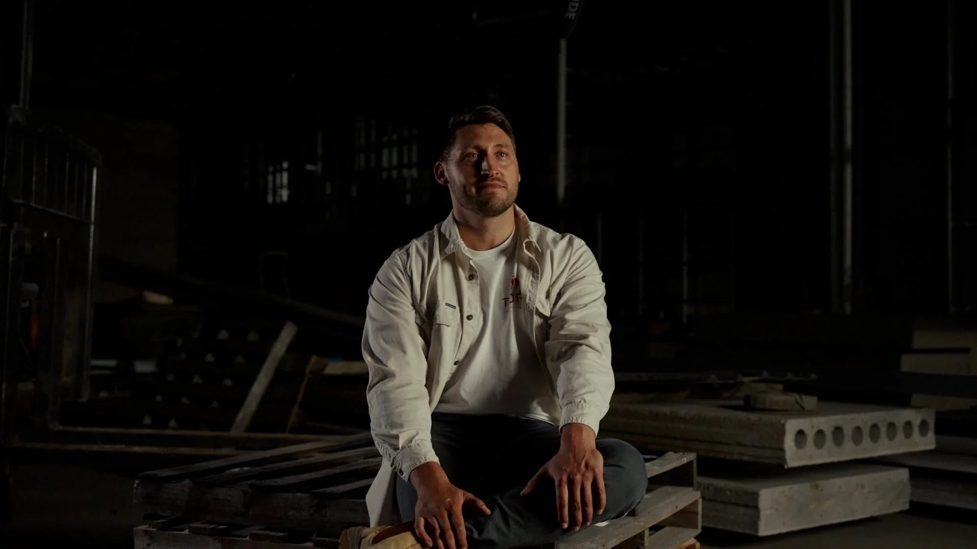 Saxon from Toro Construction Group sitting cross-legged on a wooden pallet in a dimly lit industrial or construction setting, surrounded by building materials and metal structures.