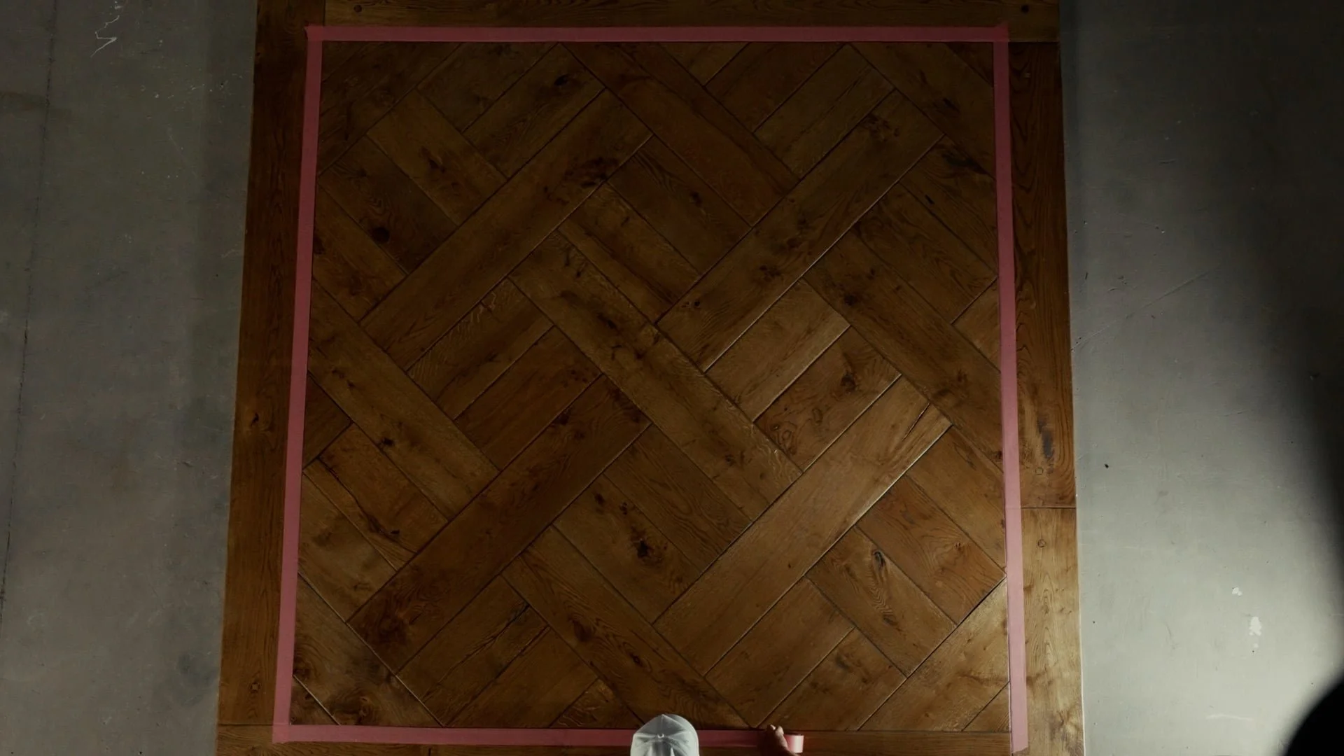 Overhead view of a square wooden parquet floor bordered by pink painter's tape, bordered by concrete flooring, with a person's foot visible at the bottom.