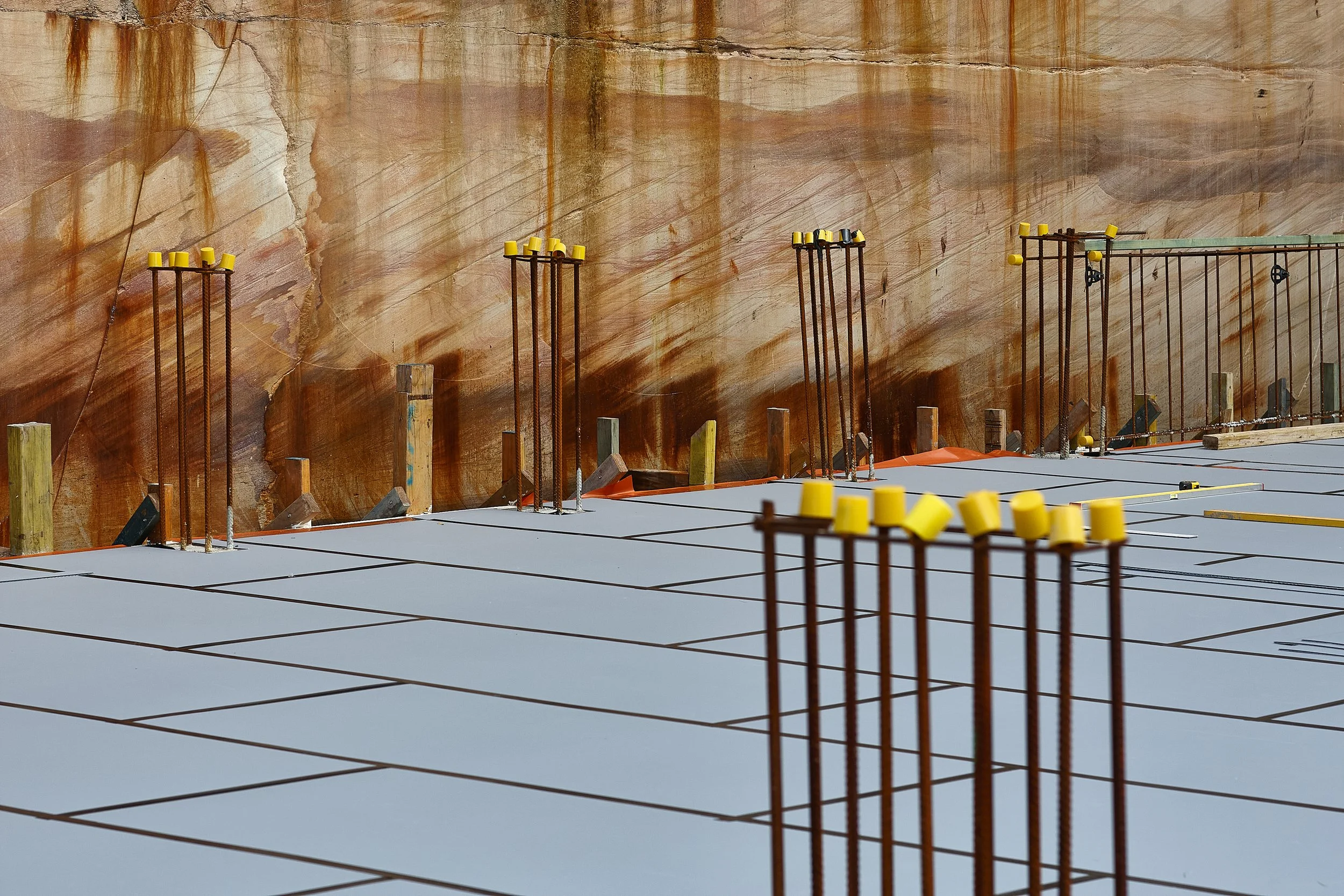 Construction site with rebar and yellow caps on a concrete slab, and a textured marble wall in the background.