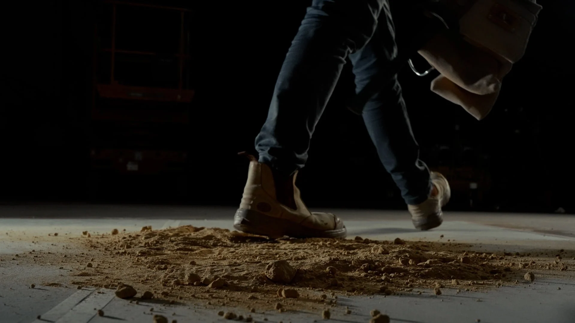 Person wearing beige boots and dark pants bending over on a dirt court, holding a brown bag with a dark background.
