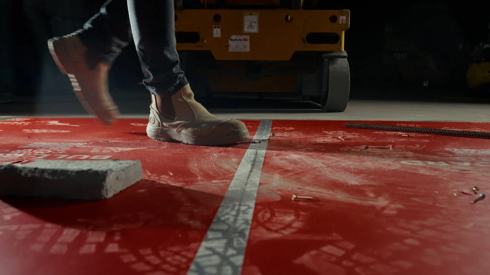 Close-up of a worker's feet and legs wearing beige safety boots and black pants, standing on a red floor with white markings, possibly in a warehouse or construction site, with a yellow industrial machine in the background.