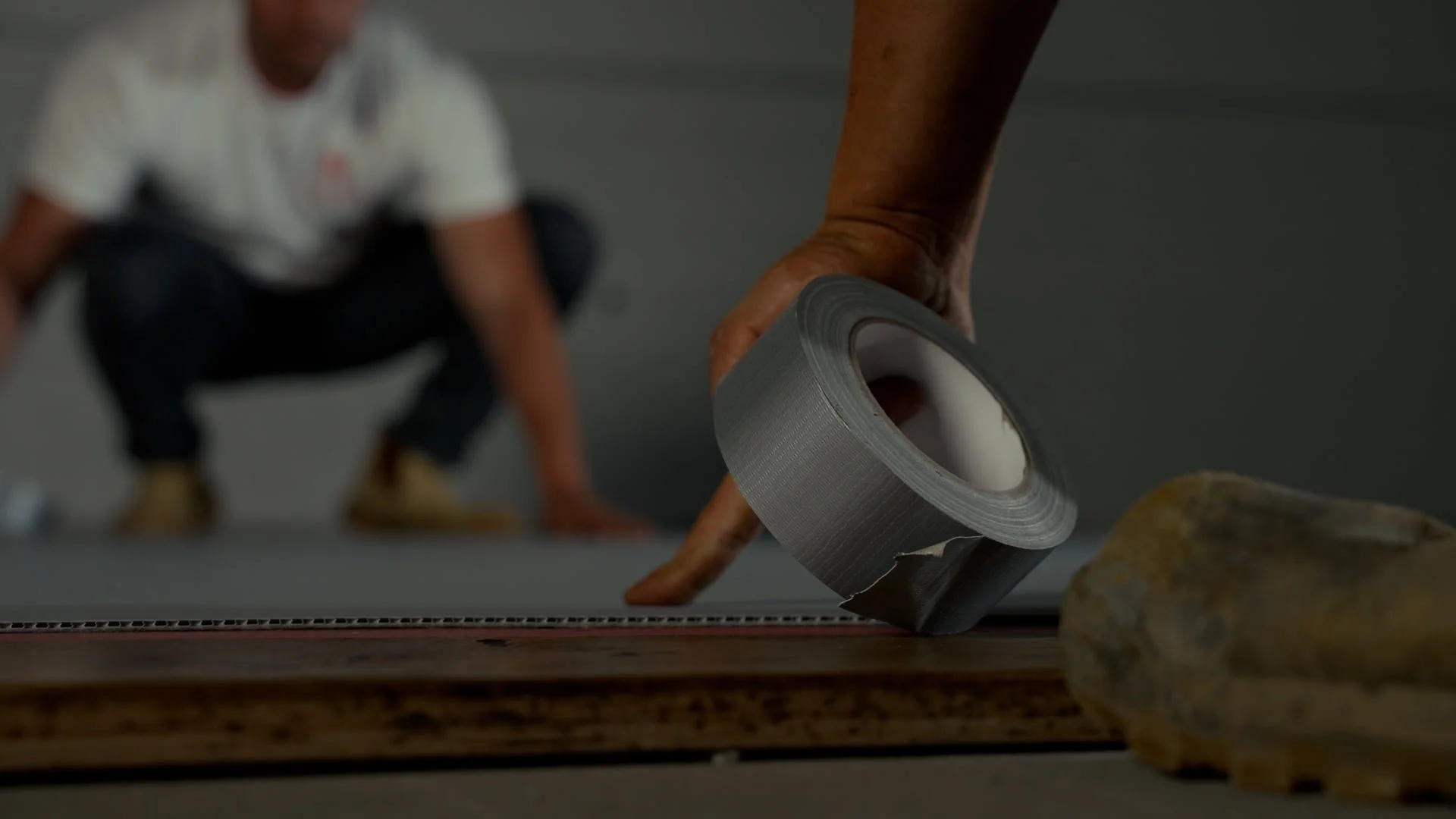 Close-up of a hand applying gray duct tape to a surface with an individual squatting in the background.