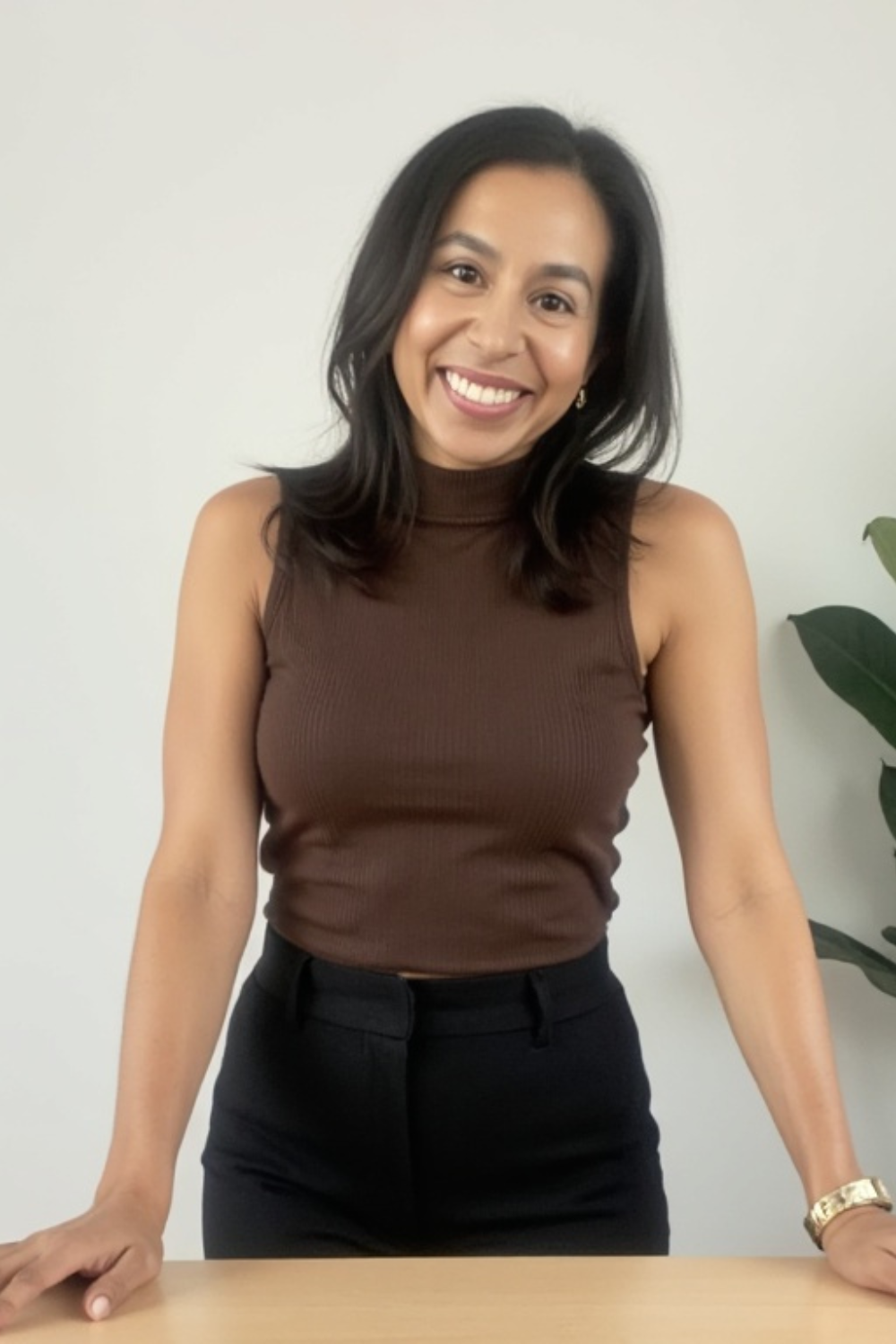 A woman with shoulder-length black hair, wearing a sleeveless brown top, smiling and leaning on a wooden table.