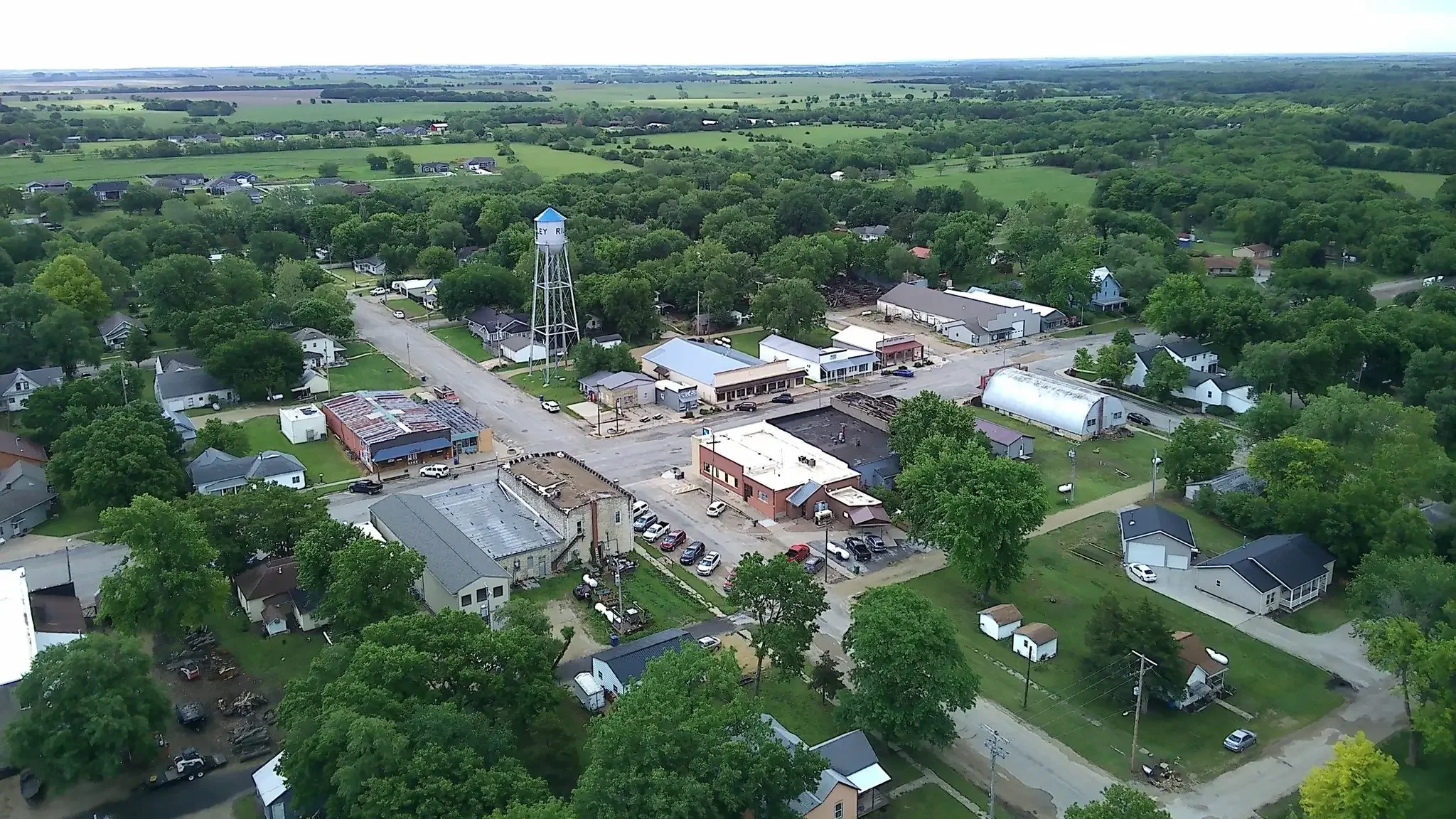 Aerial view of a small town with residential houses, a water tower, commercial buildings, and a green landscape in the background.