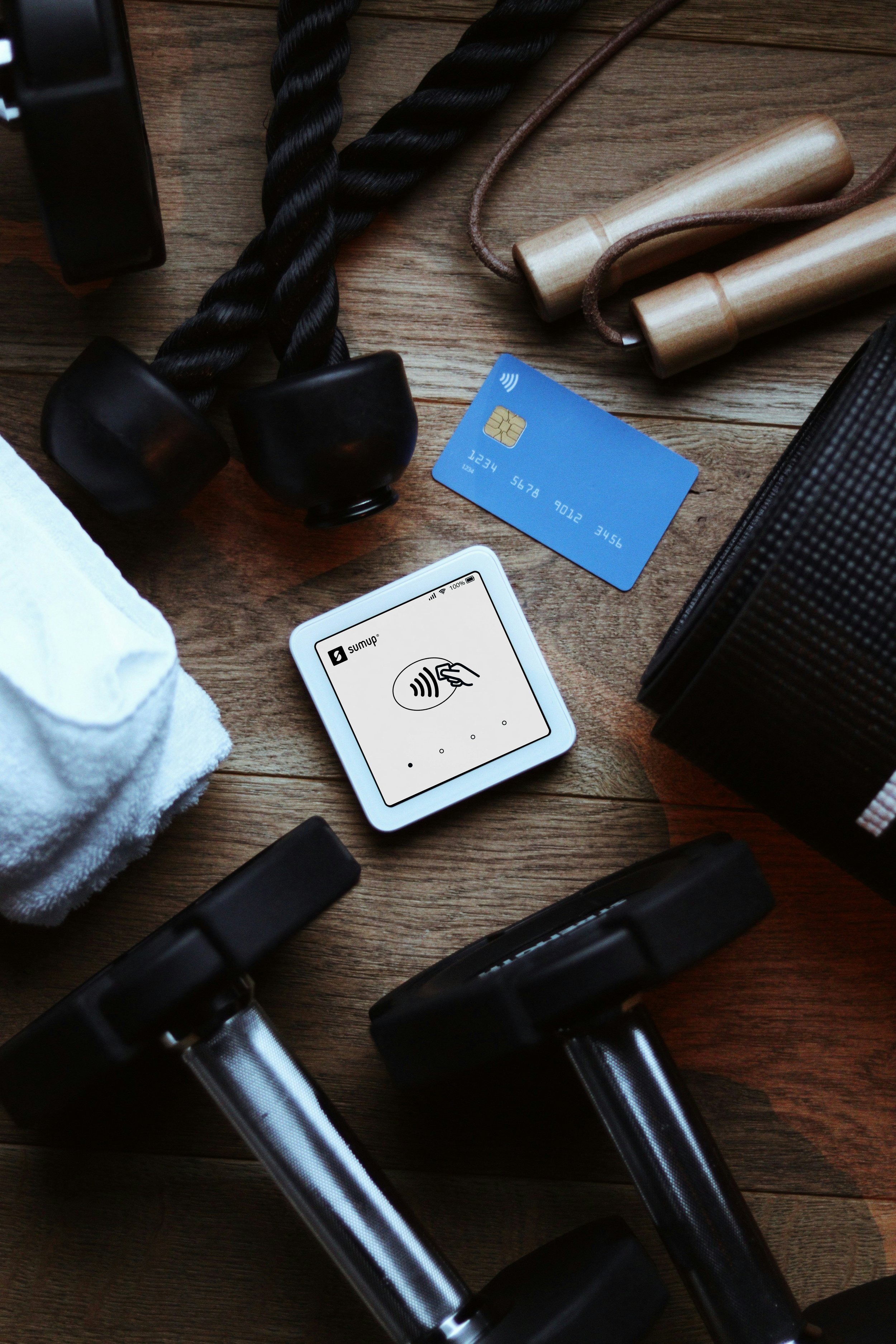 Fitness equipment including a pair of dumbbells, a jump rope, a white towel, a blue contactless payment card, and a digital payment device on a wooden floor.