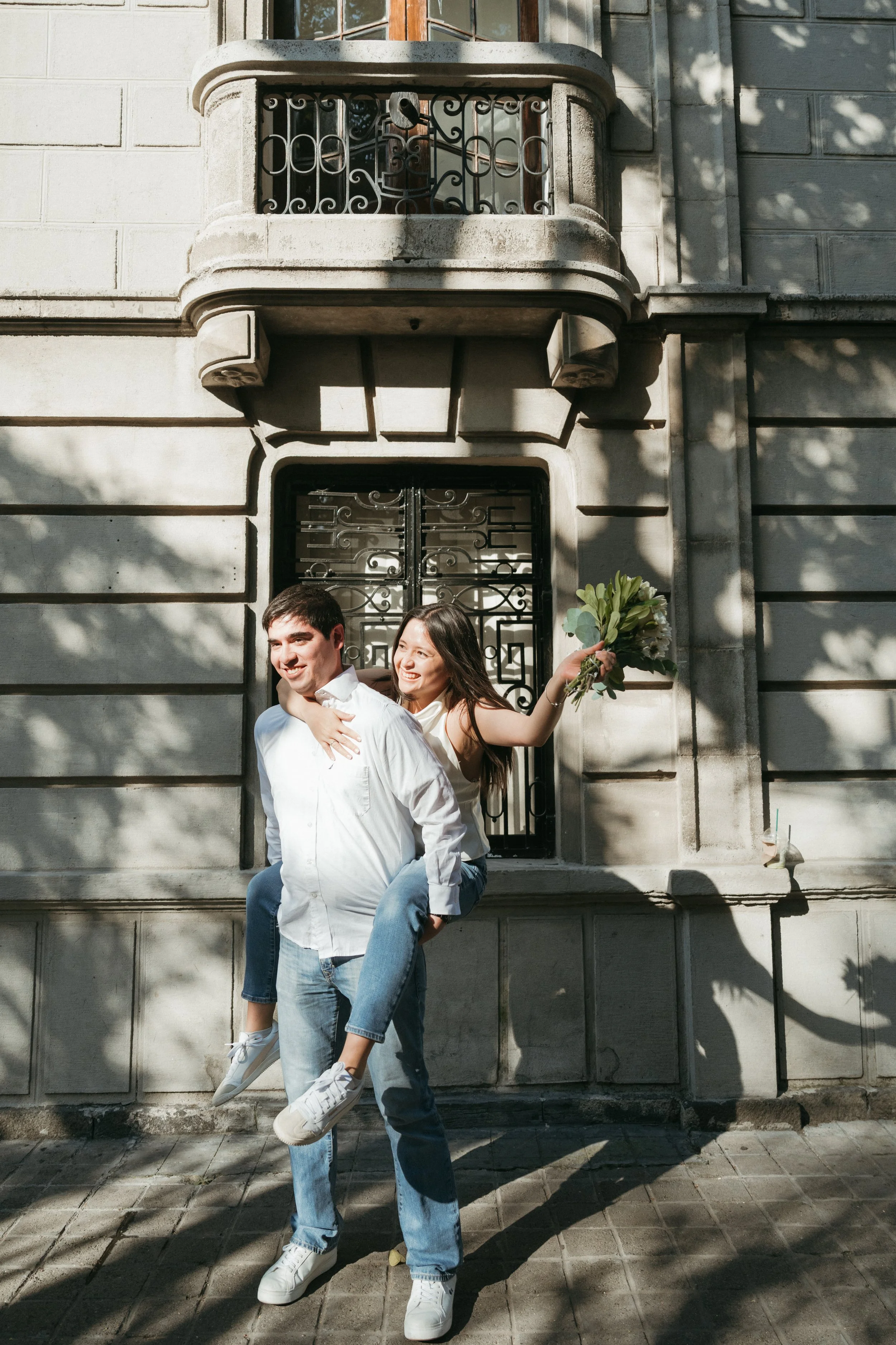 A smiling couple outdoors, with the woman holding a bouquet of flowers on the man's back, standing in front of an ornate building facade with shadows of trees.