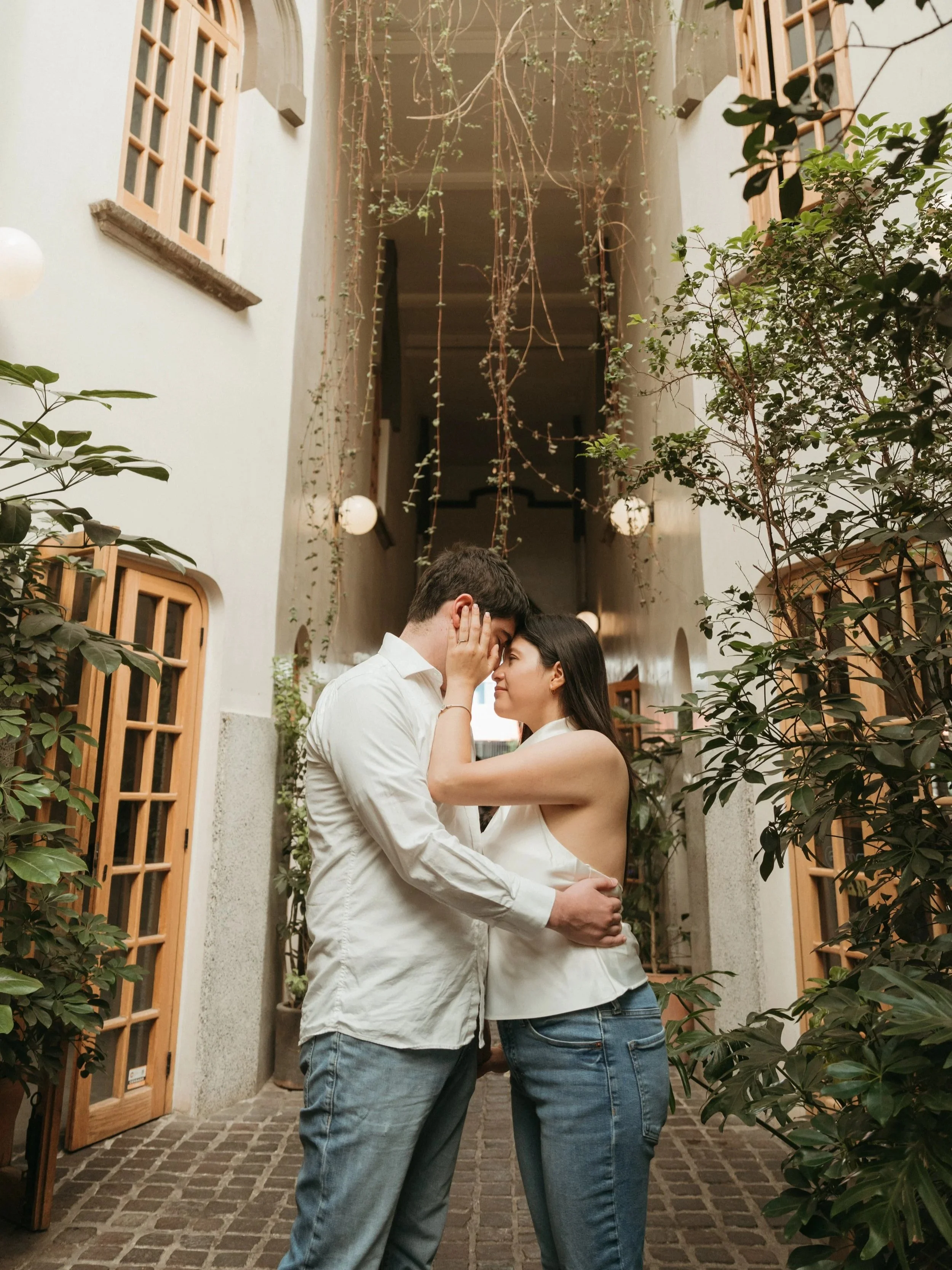 A couple is embracing and gently touching foreheads in a cozy, plant-filled outdoor corridor.