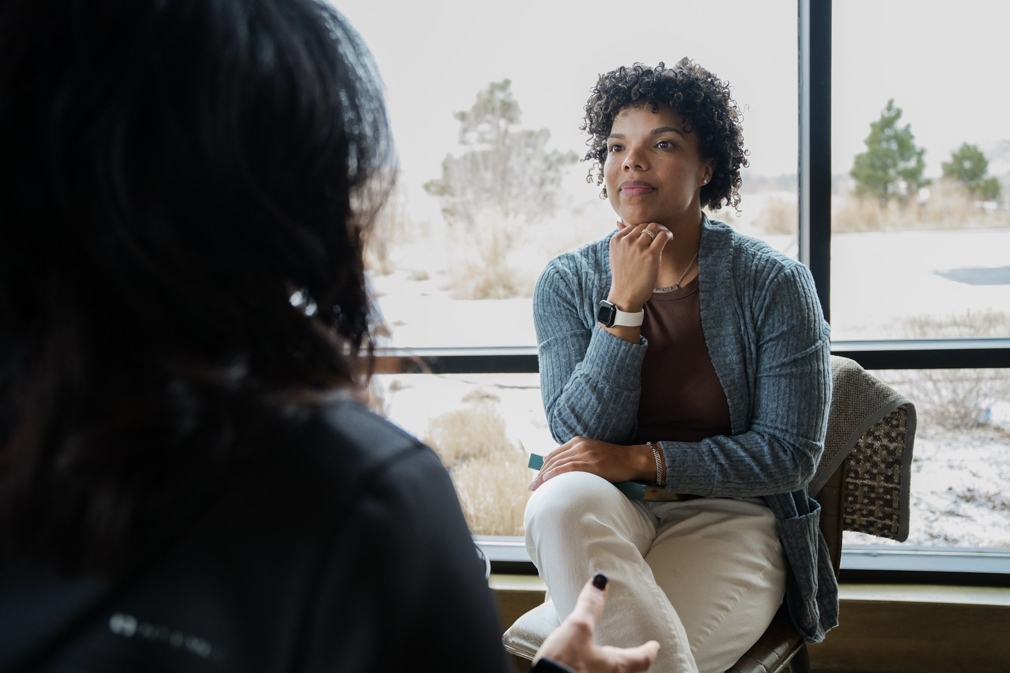 Two women having a conversation indoors near a large window with a snowy landscape outside. One woman is facing the camera, sitting on a chair, with curly hair, wearing a gray cardigan and brown top. The other woman, with dark hair, is seen from behind, gesturing with her hand.