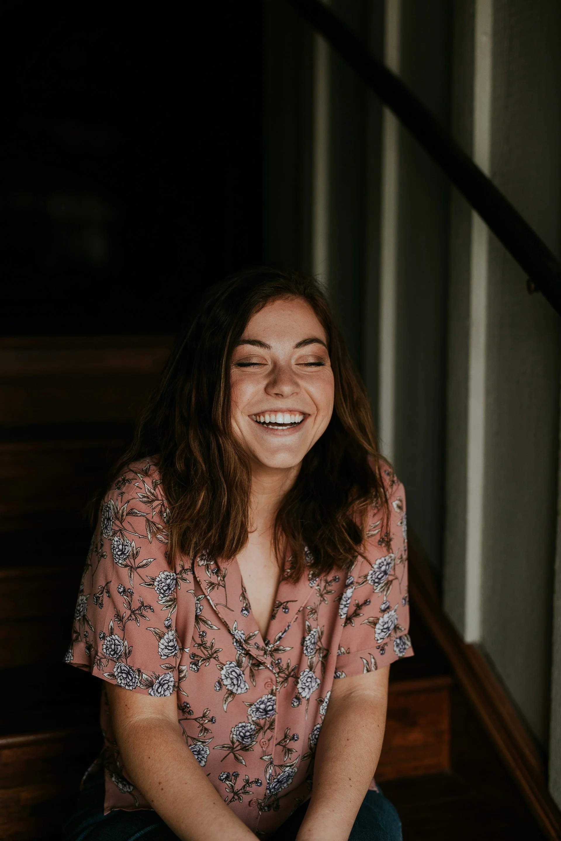 A young woman with brown hair and a floral pink blouse sitting on stairs, smiling with eyes closed.