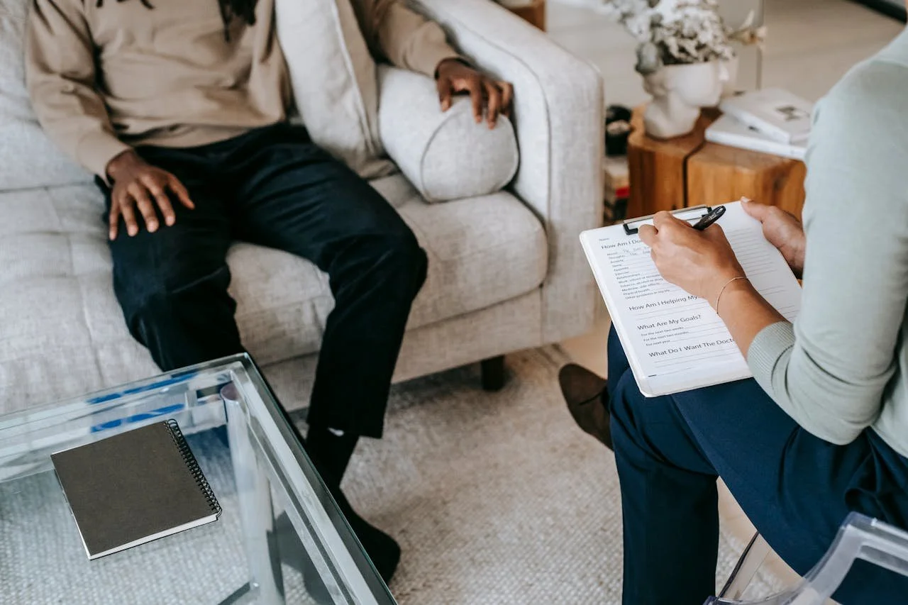 A person in a light-colored shirt taking notes on a clipboard during a therapy or counseling session with a patient sitting on a couch.