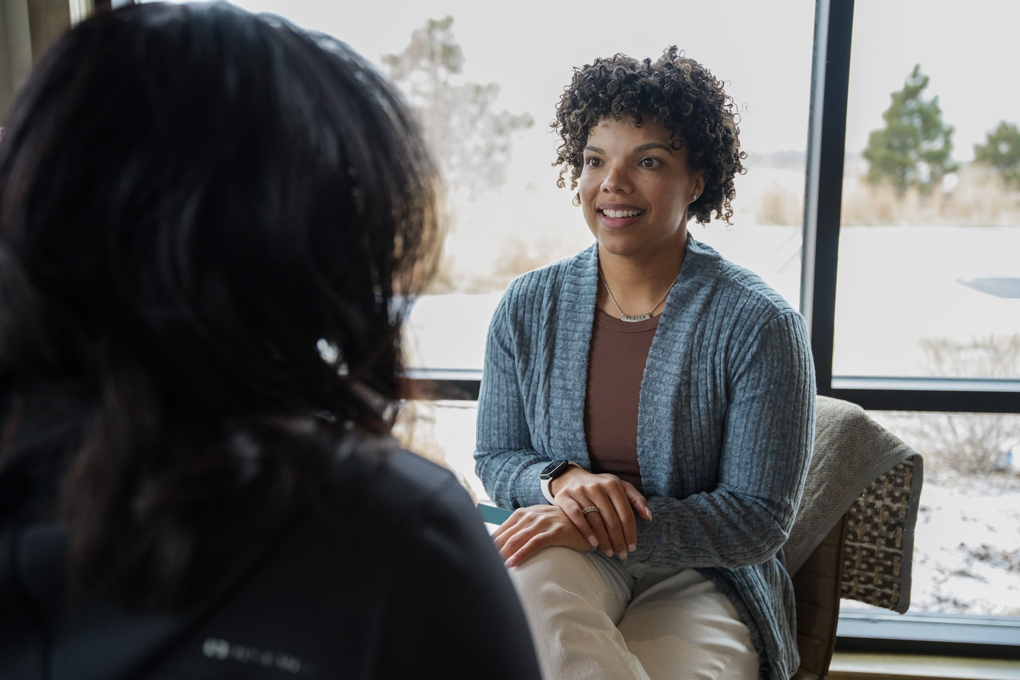 Two women having a conversation in a bright room with large windows, one with curly hair wearing a blue cardigan, and the other with dark hair.
