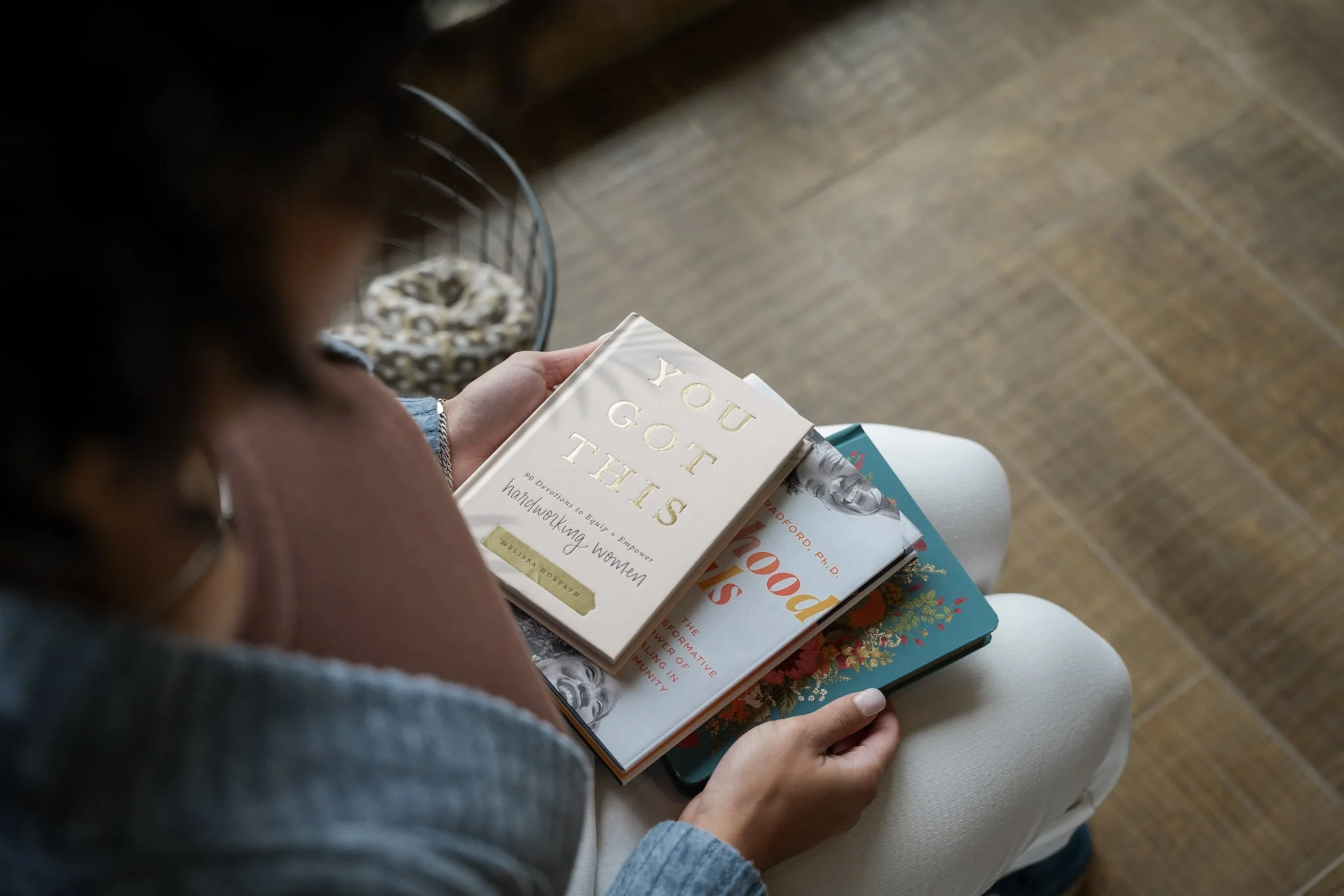 A person sitting in a white chair reading three books, with a basket of dog toys nearby. The book on top is titled 'You Got This' by Melissa Orlady.