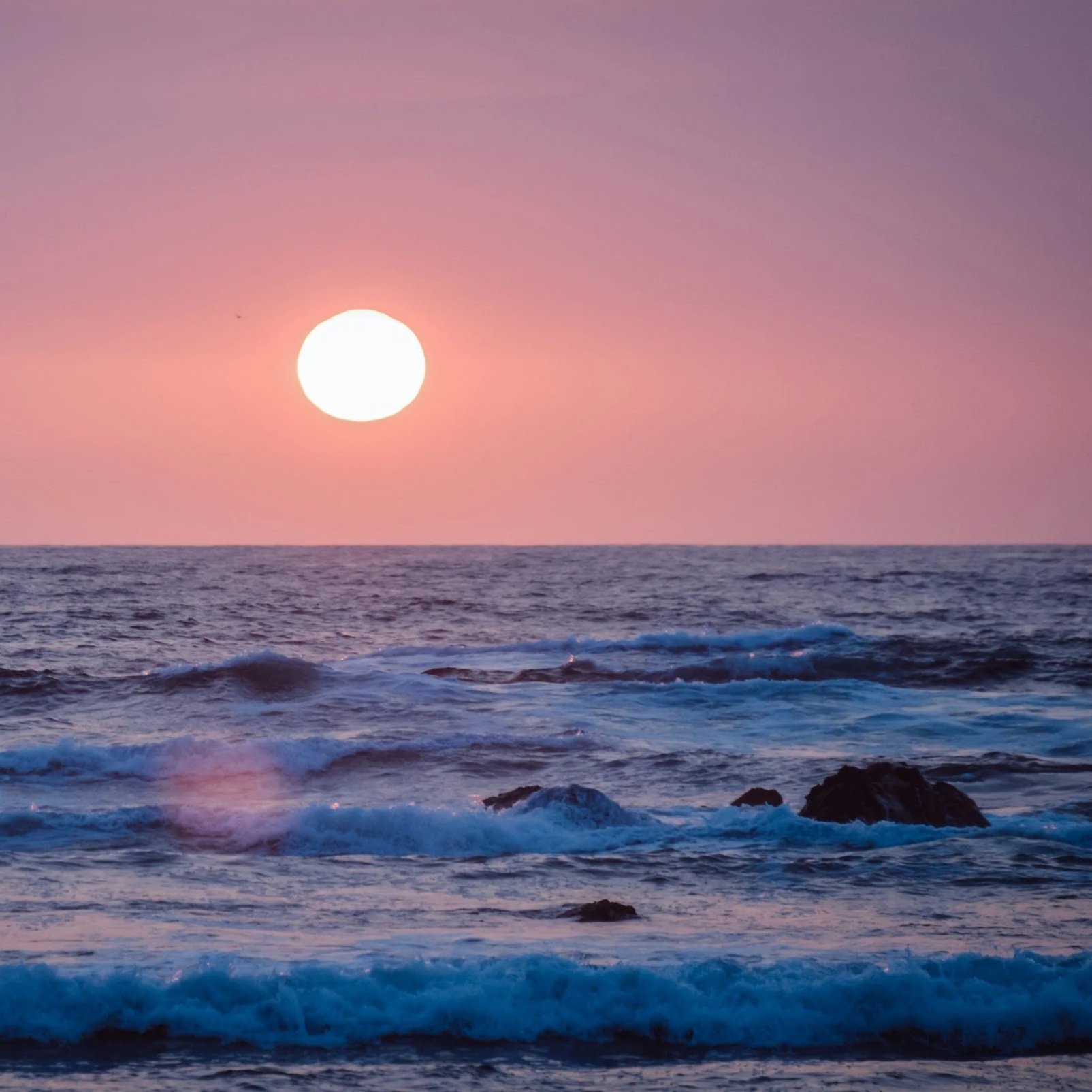 Sunset over the ocean with pink and purple clouds and waves crashing on rocks.