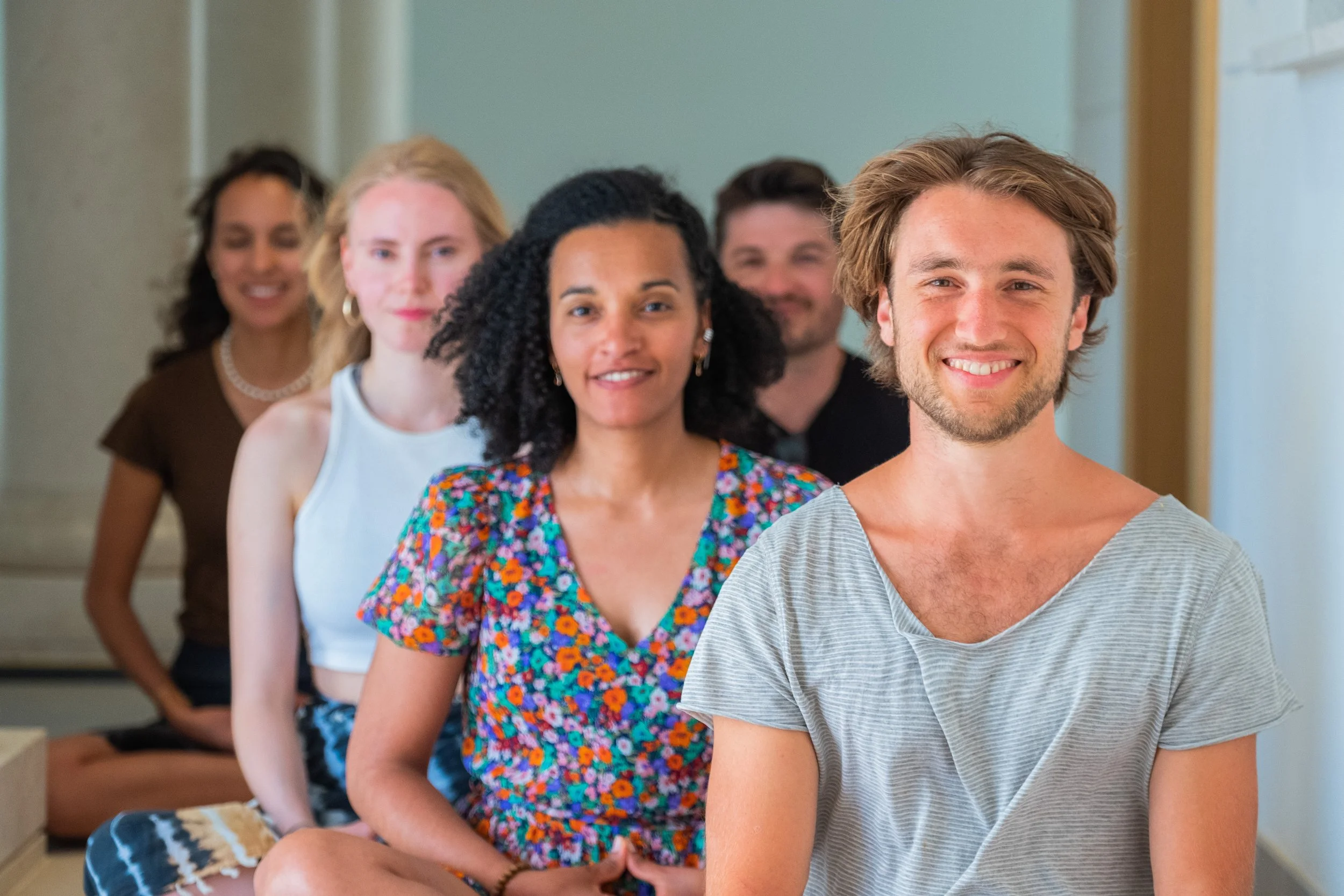 Group of six diverse smiling people sitting in a line indoors.
