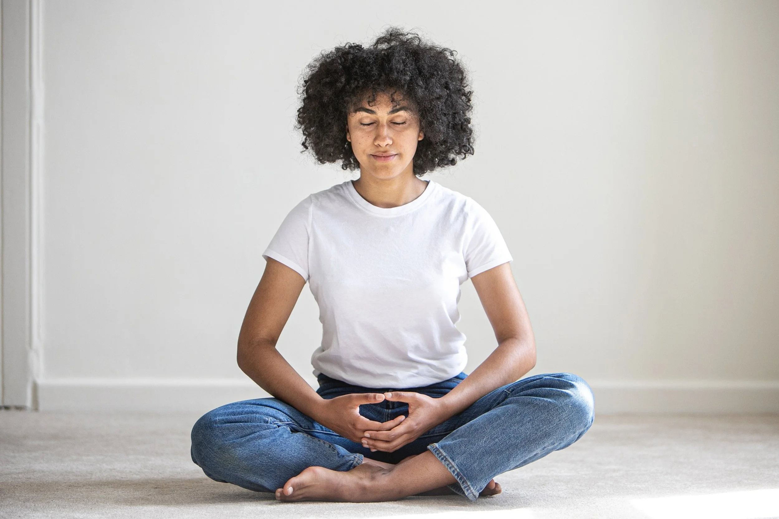 Image of peaceful Kadampa meditator seated on the floor in meditation posture with white tee shirt, blue jeans and bare feet and voluminous curly hair around her head and ears