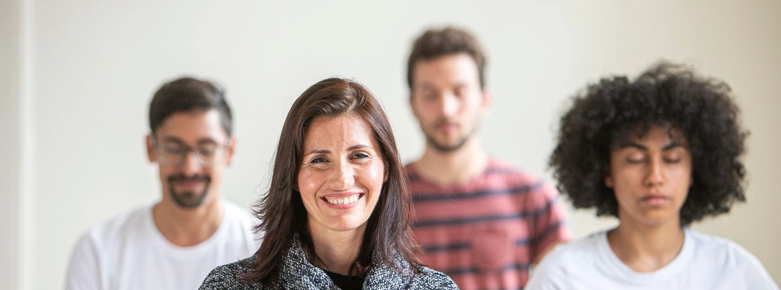 Group of four diverse adults seated meditating together, with one woman with long hair smiling in the foreground.