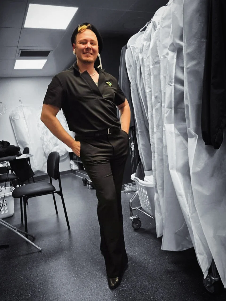 image black and white of founder and owner in a black shirt and pants hands in pocket. Broche on lapel and a hat. Person is in color. background reads as dressing room.
