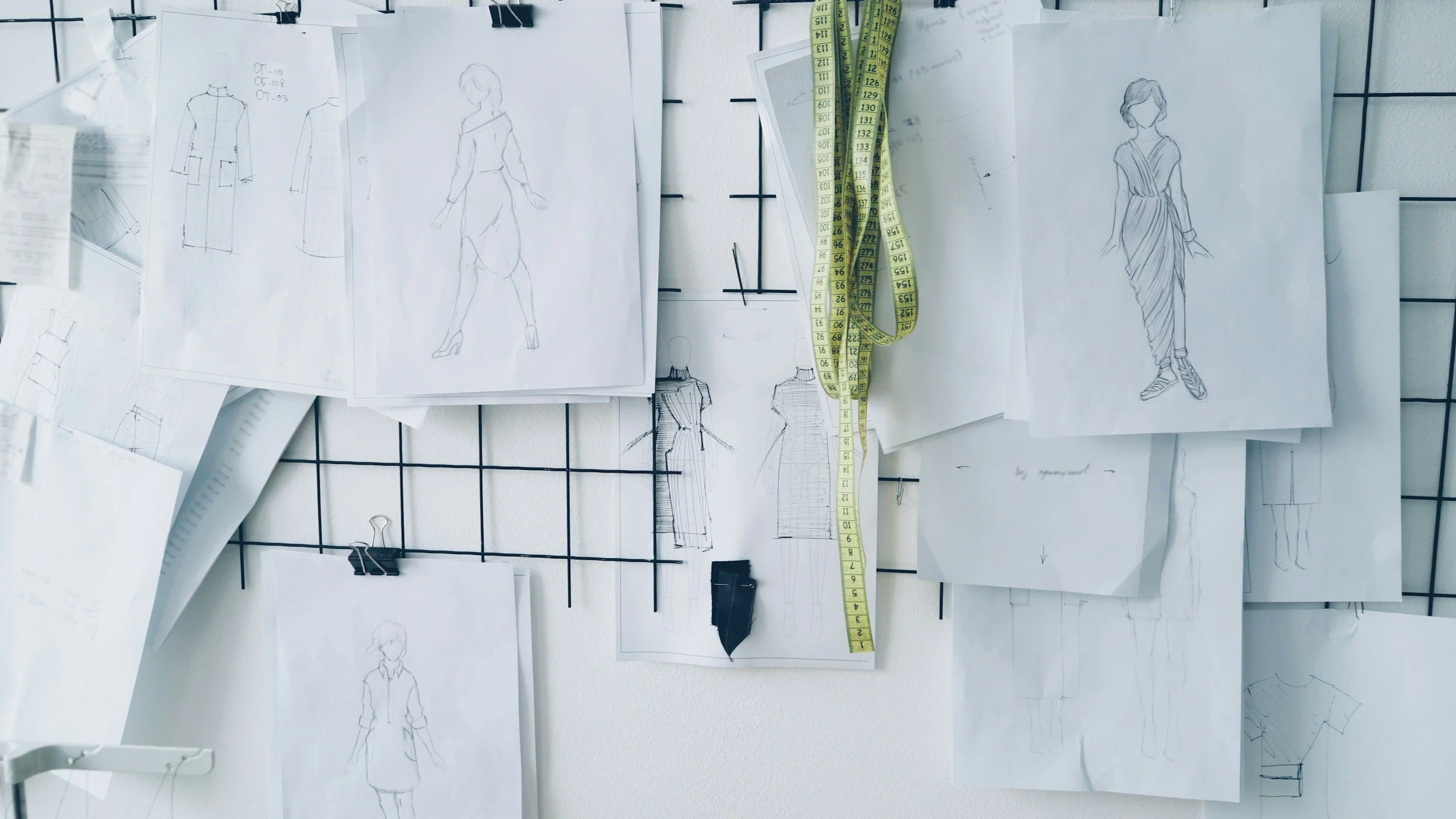 Sketches on white paper of three different dresses hanging on a black wire wall display and a large yellow tailors measuring tape hanging off center