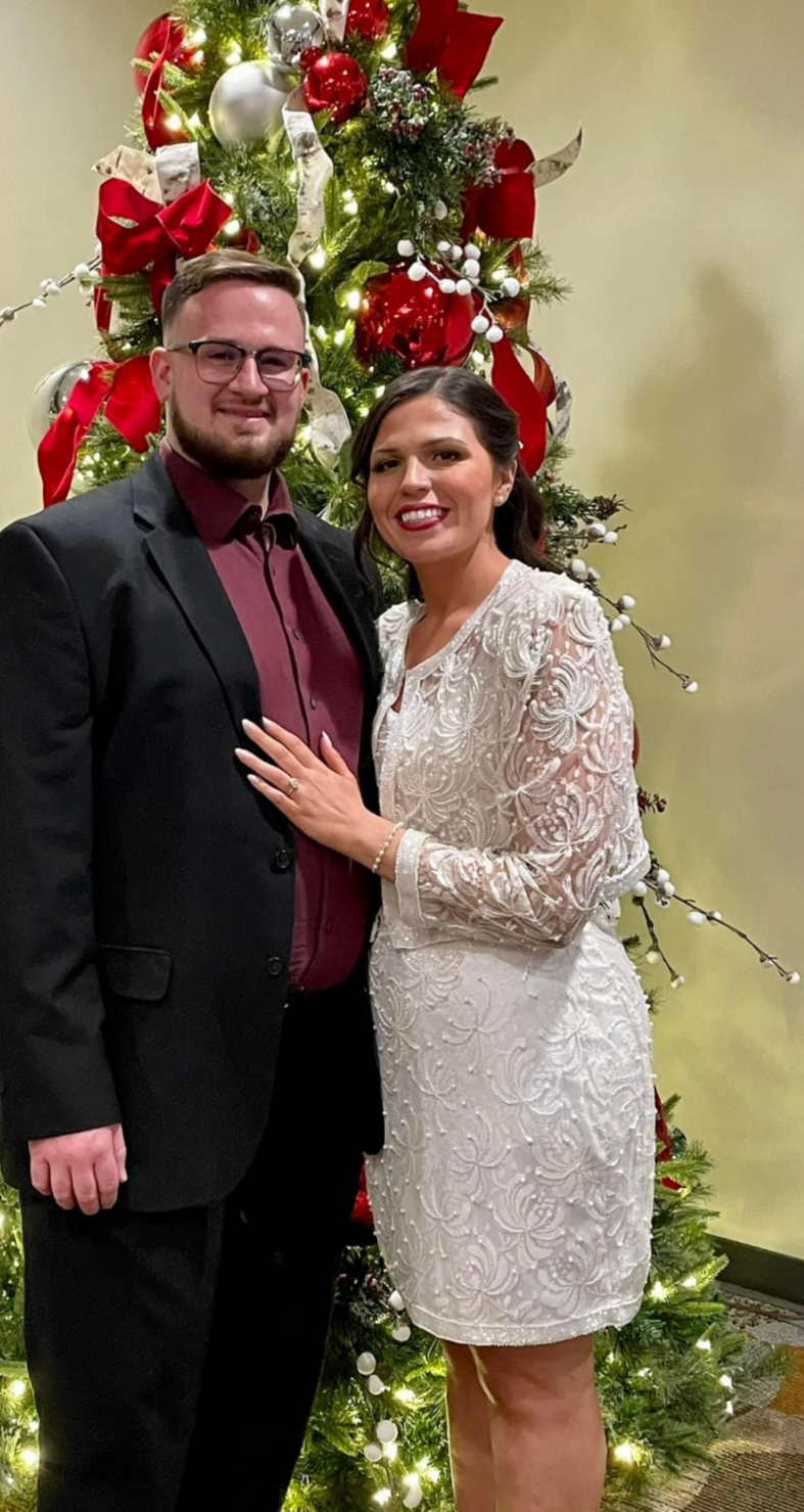 Woman in a beaded dress with bolero in front of a Christmas tree with her soon to be husband in a dark suit and bowtie