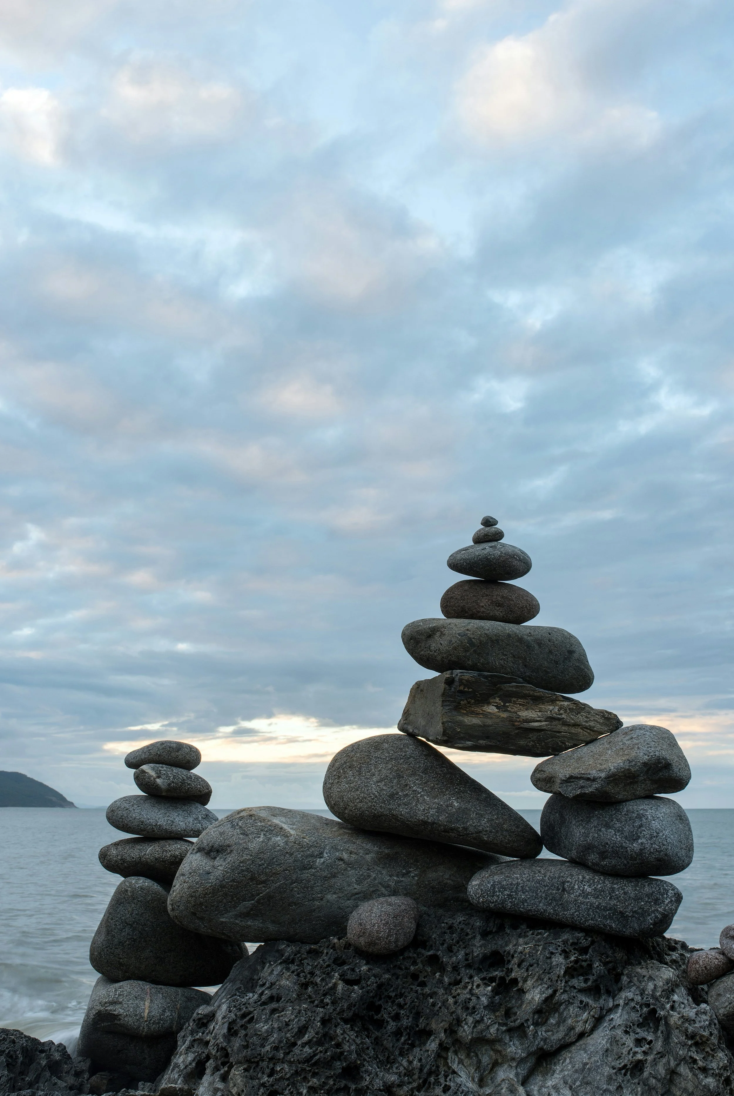 Stacked rocks on a rocky shoreline with a cloudy sky and distant land in the background.