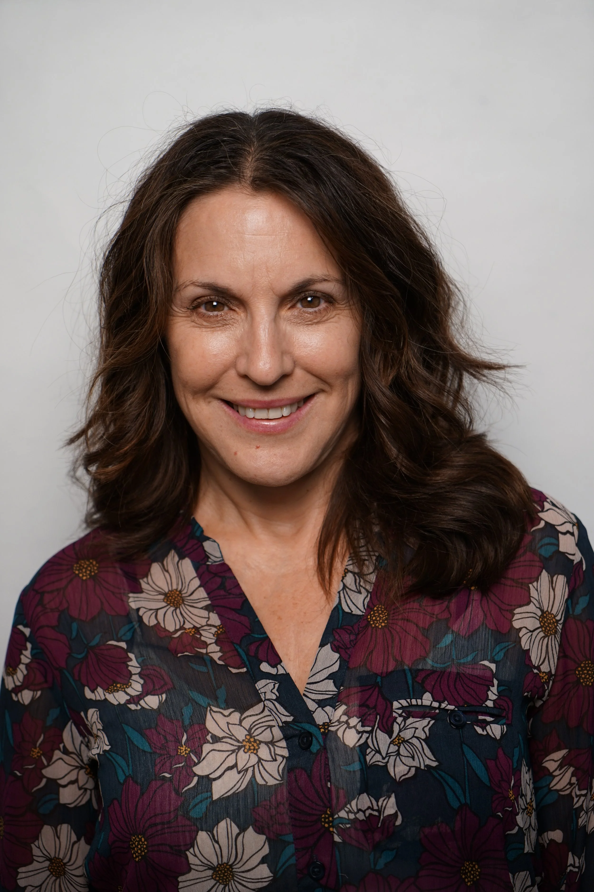 A woman with wavy brown hair, smiling and wearing a floral blouse, standing against a plain light gray background.