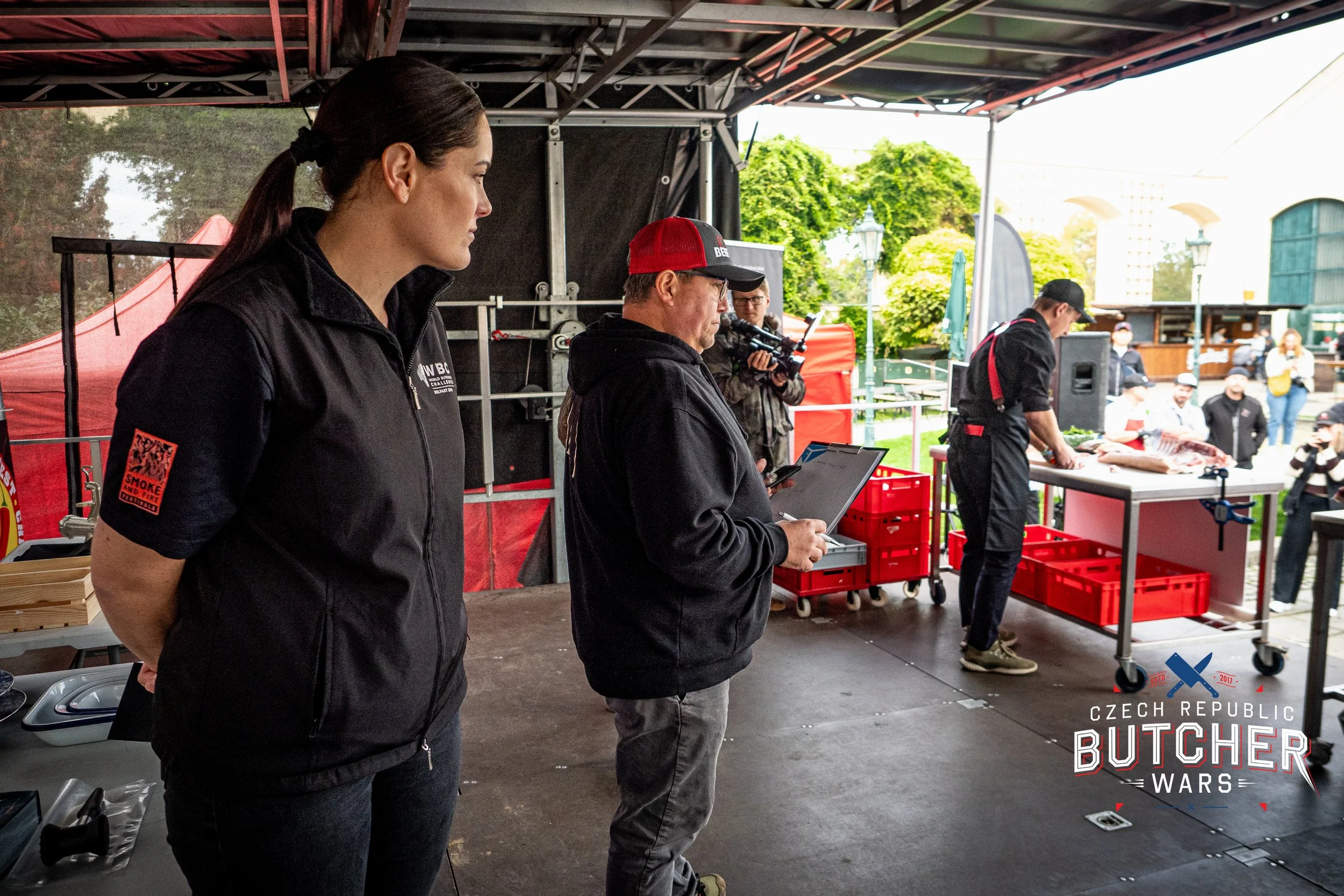 People preparing meat at an outdoor event during the Czech Republic Butcher Wars, with people watching, some taking photos, and a woman in the foreground wearing a black jacket with red patch on the sleeve.