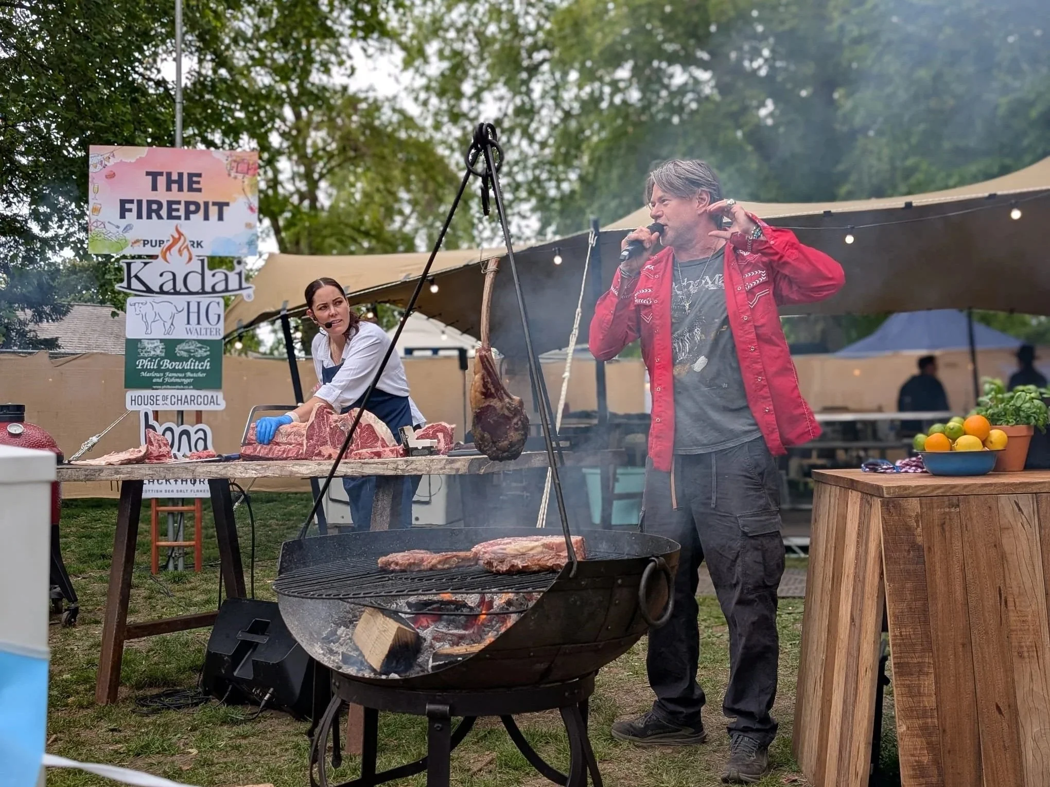 A man with grey hair wearing a red jacket and grey pants is speaking into a microphone at an outdoor barbecue event. A woman wearing a white shirt and apron is preparing meat on a table with a hand saw. 