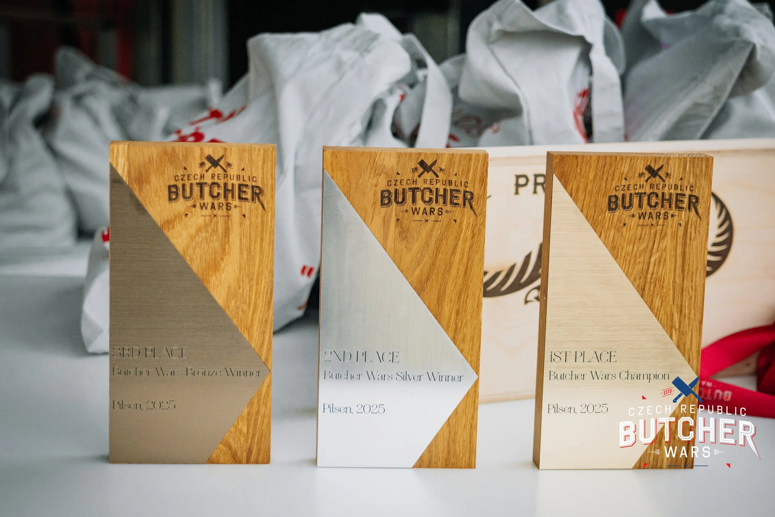 Three wooden trophies with geometric design, each with the 'Czech Republic Butcher Wars' logo on top, indicating 3rd, 2nd, and 1st place awards, with the year 2025 and the word ‘Pilsen’ at the bottom. The trophies are arranged in a row on a white sur
