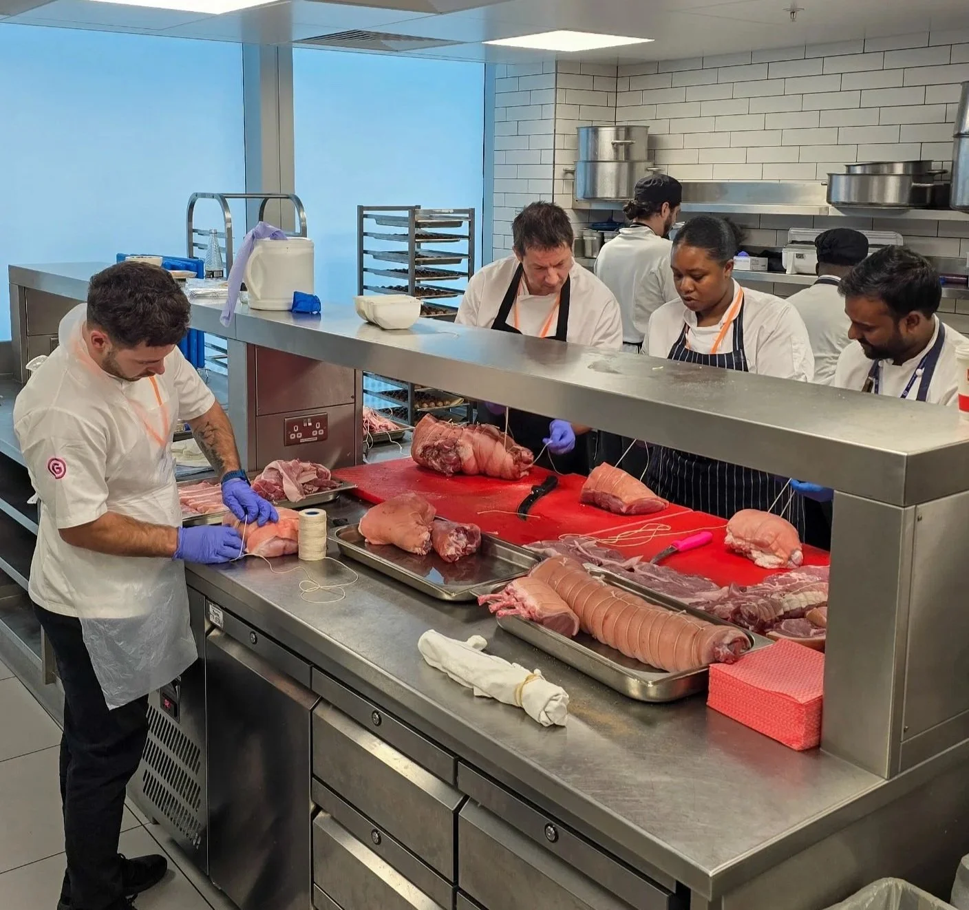 Chefs butchering pork under the guidance of Alayna Empson in a professional kitchen with stainless steel equipment and white tiled walls.