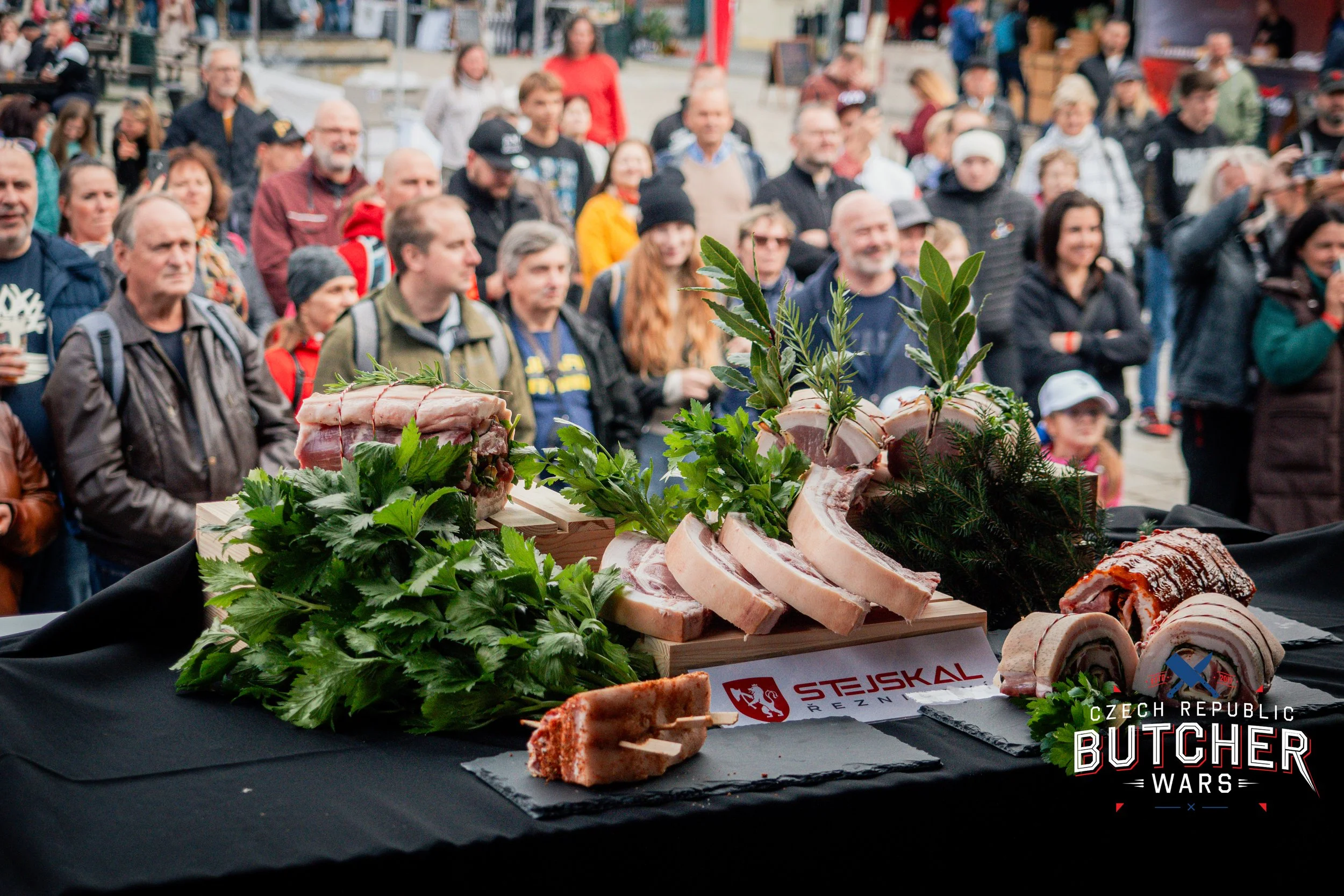 Display of raw meats, including ribs and pork belly, garnished with fresh green herbs on a table at a public event, with a crowd of people watching in the background.