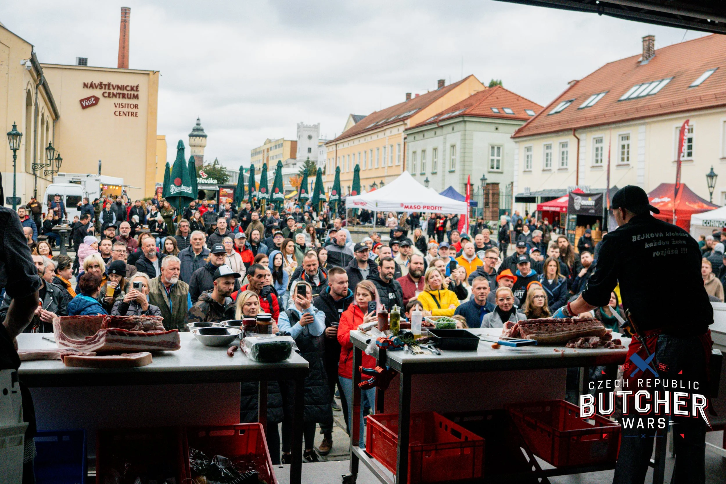 A large crowd gathered outdoors at a meat grilling event in Czech Republic, with a butcher demonstrating cutting meat on a table. People are watching and taking photos, and tents and buildings are visible in the background.